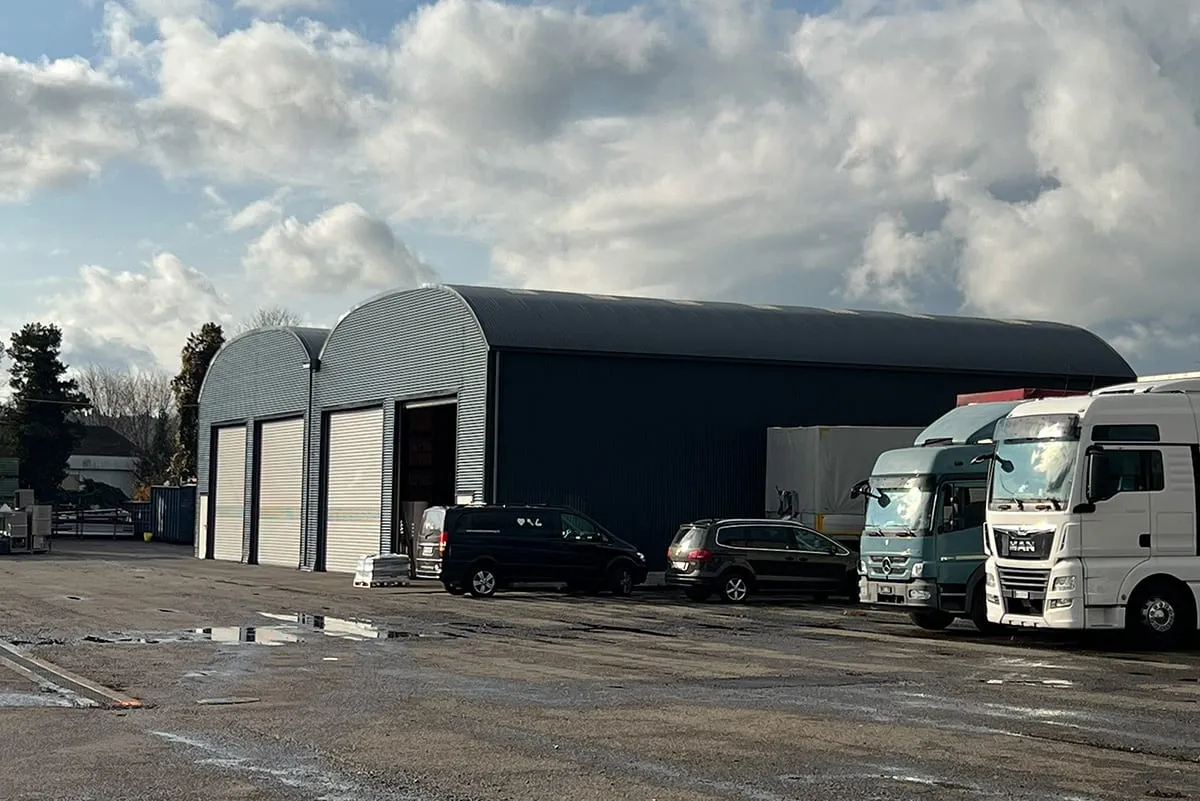 Dark grey storage hall with an arched roof and three roller doors, vans and a lorry parked in front