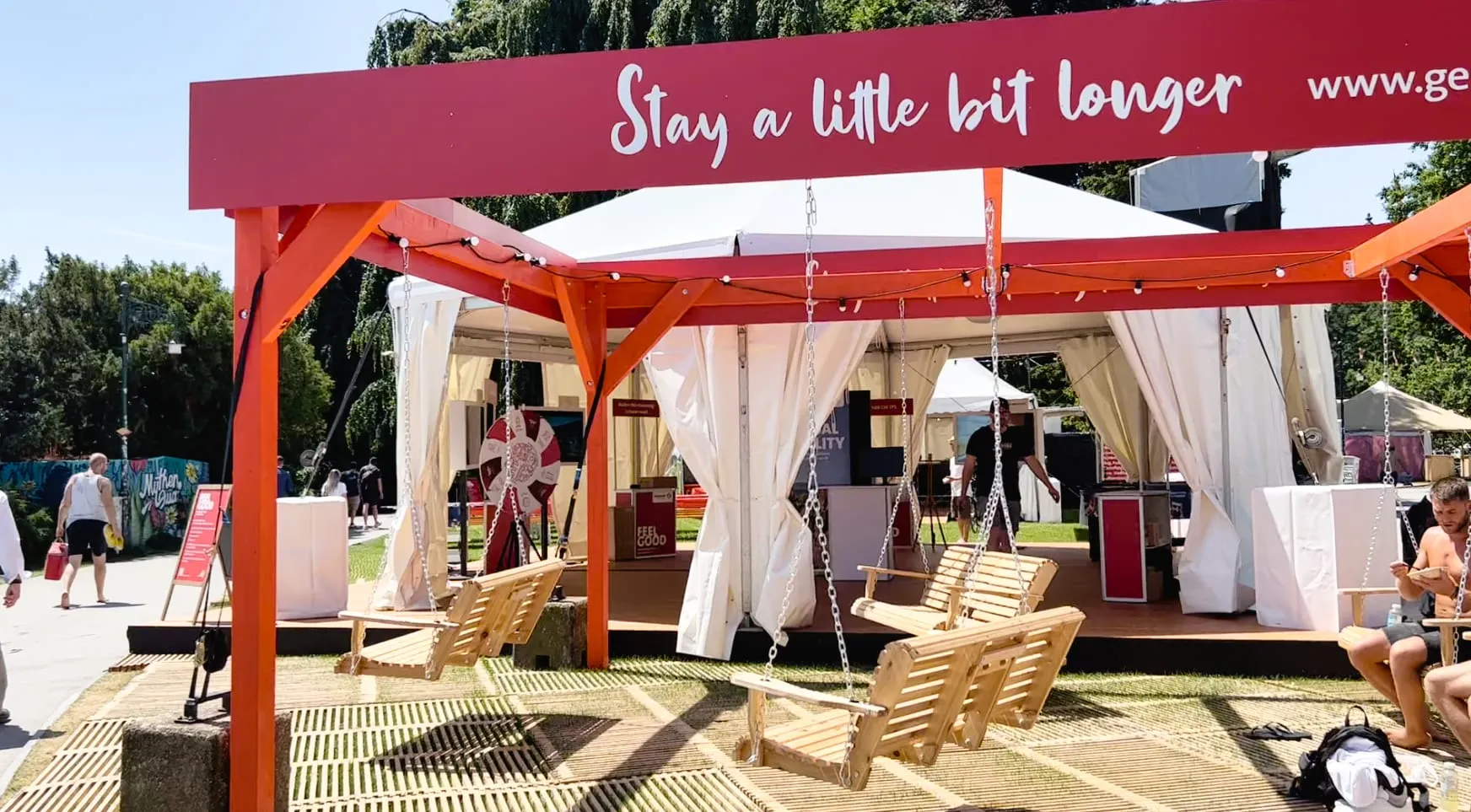 Red pergola with «Stay a little bit longer» lettering and wooden swings in front of the white DZT tent with a Glücksrad