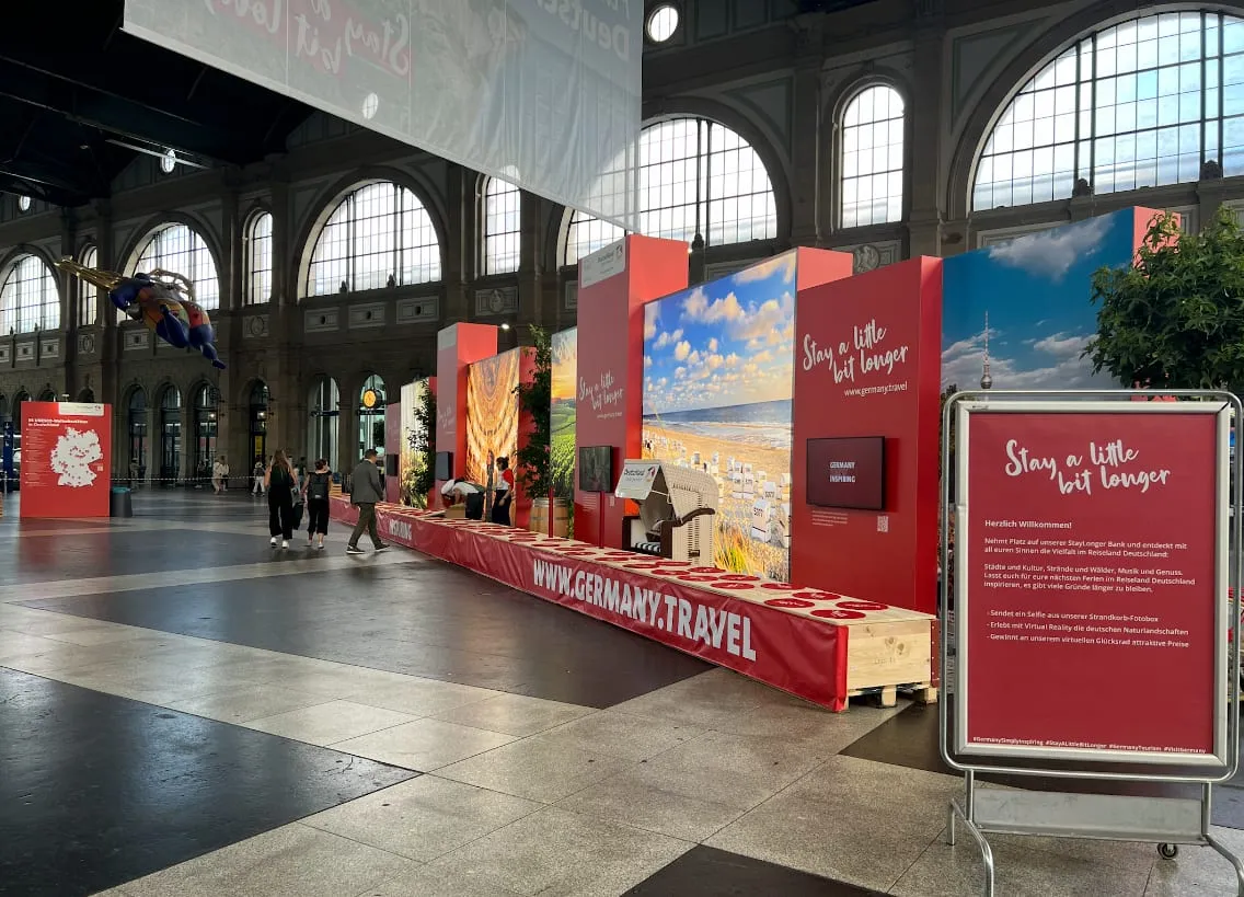 Lange Holzbank mit Banner www.germany.travel vor roten Wänden und Strandkorb in der Bahnhofshalle