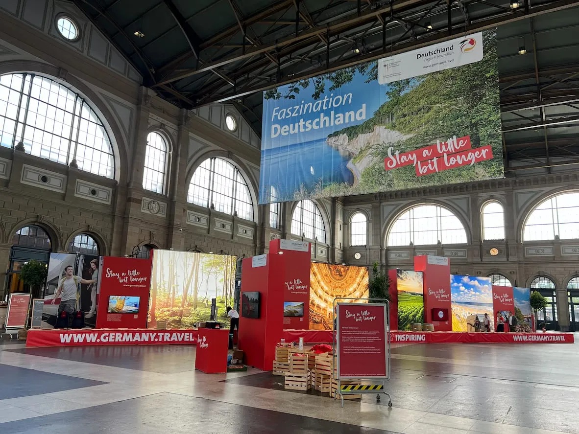 Red exhibition walls with landscape photos and an XXL «Faszination Deutschland» banner in the hall of Zürich main station