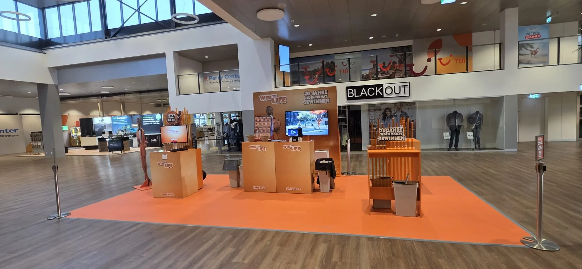 Orange Win for Life stand with a glitter counter, screen, «20 Jahre jeden Monat gewinnen» backdrop and orange carpet in a shopping centre