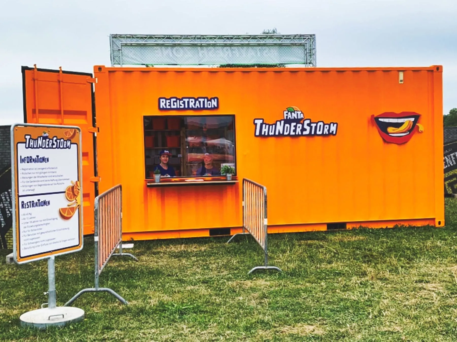 Orange Fanta Thunderstorm container with a registration desk, crowd barriers and an information board on a meadow