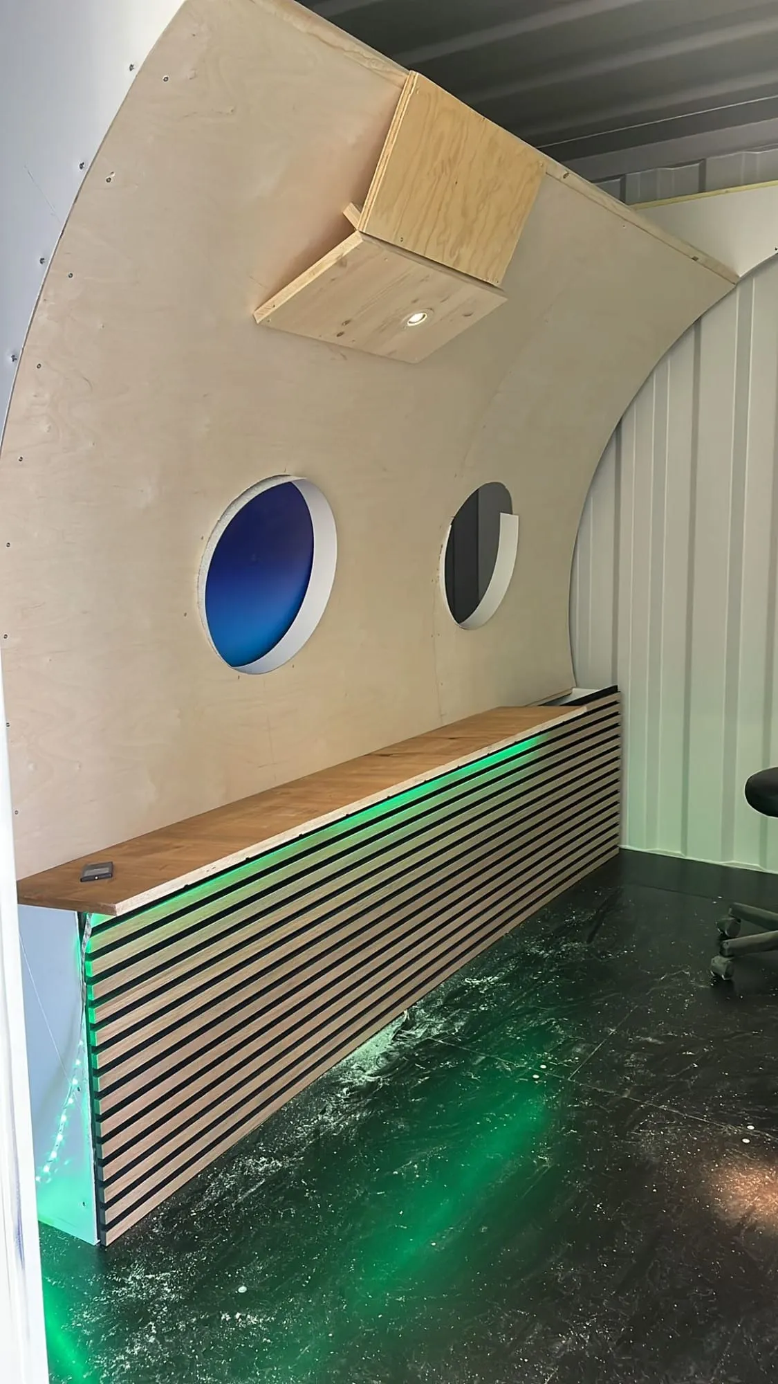 Rohbau des Jets im Container: gewölbte Holzwand mit zwei Bullaugen und Lattentheke mit grünem LED-Licht