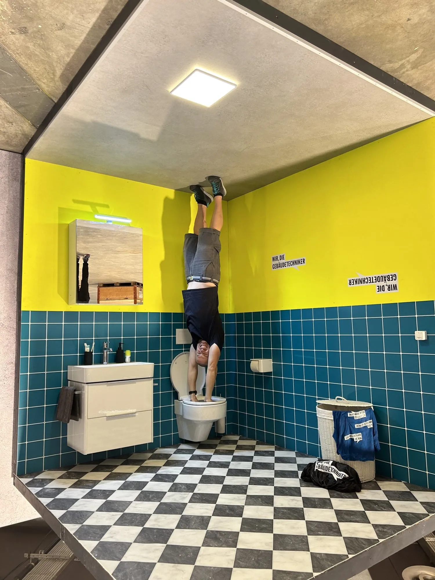 Mann macht Handstand auf dem WC im fertigen Upside-Down-Bad mit Schildern «Wir, die Gebäudetechniker»