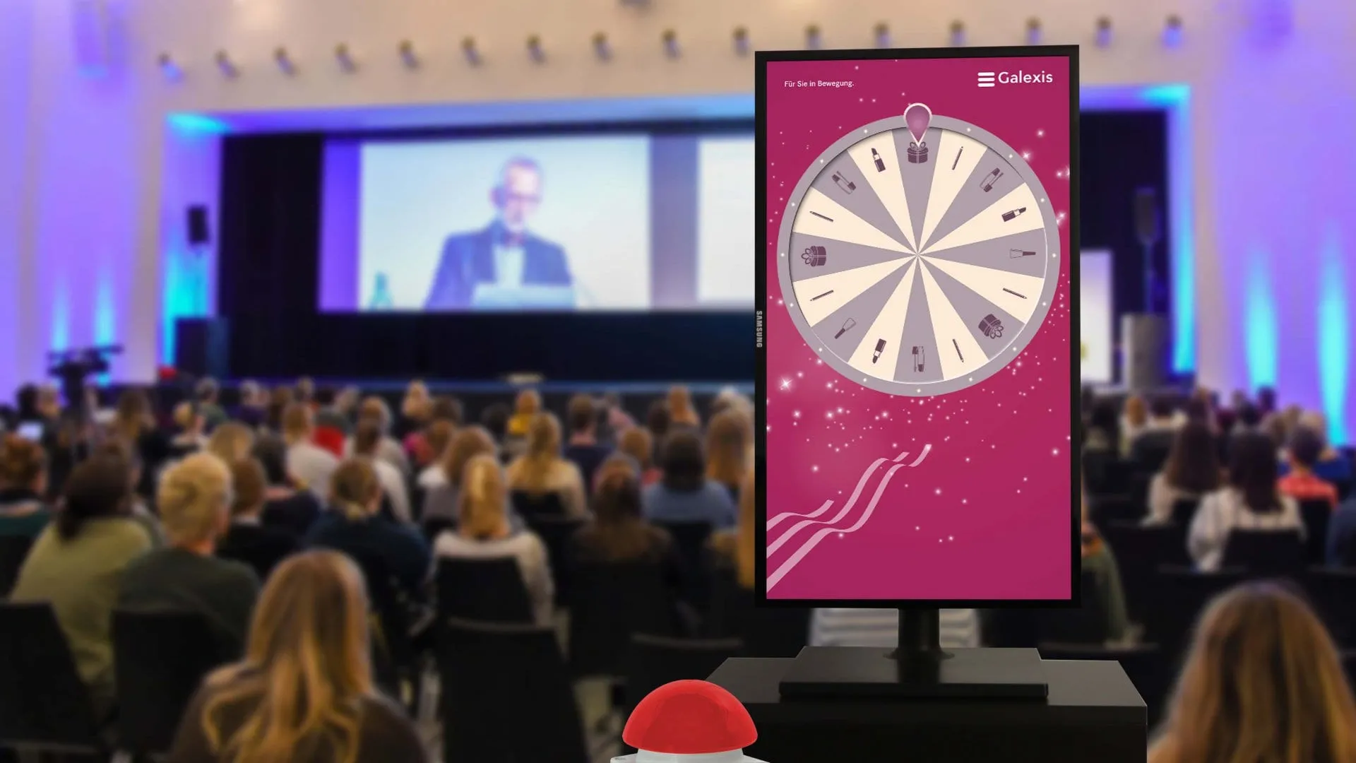 Portrait screen with the pink Galexis Glücksrad and a red buzzer in front of a congress hall with an audience