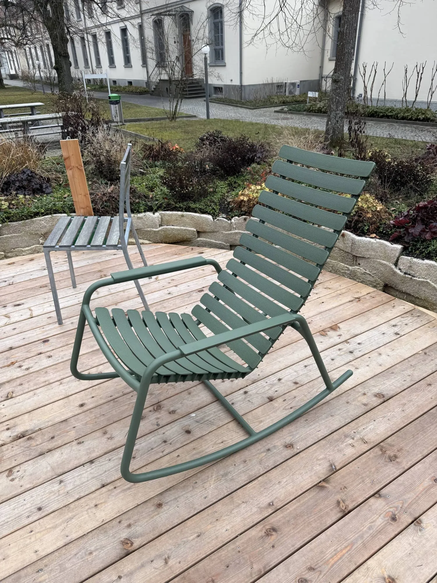 Grüner Schaukelstuhl und Metallstuhl auf dem Holzdeck des Blumenrings vor Klinikgebäuden