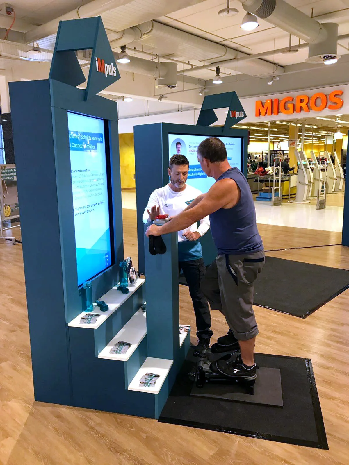 Mann auf einem Fitness-Stepper vor dem iMpuls-Glücksrad-Screen, daneben ein Promotor im Migros-Laden