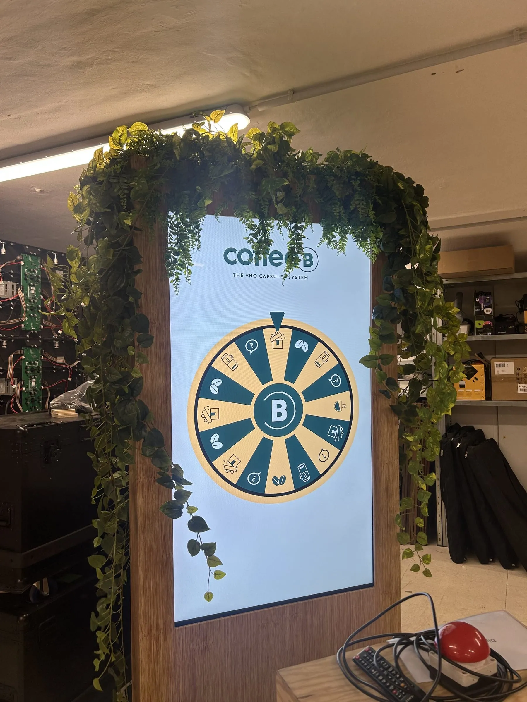 Screen mit Coffee-B-Glücksrad in Grün und Beige, eingerahmt von Holz und hängenden Kunstpflanzen, in der Werkstatt
