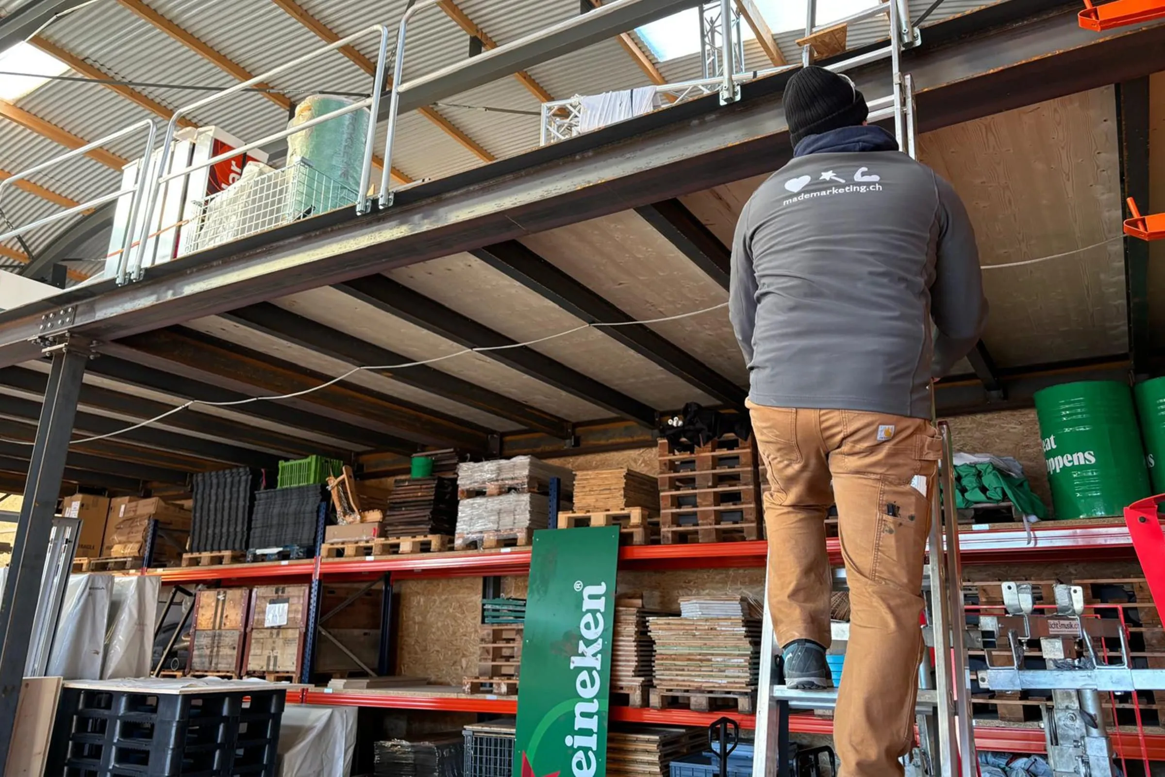 Man in a made Marketing jacket on a ladder under the mezzanine floor of the storage hall, shelves with pallets
