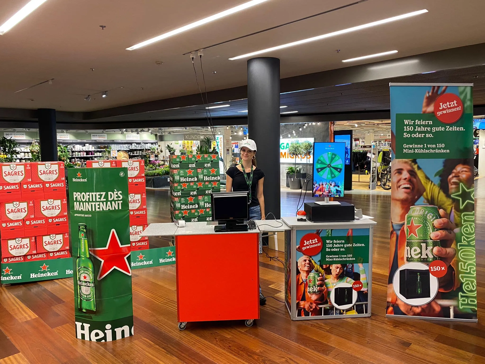 Heineken promotional stand in Migros with beer crates, a red counter, digital Glücksrad screen, red buzzer and «150 Jahre» banner