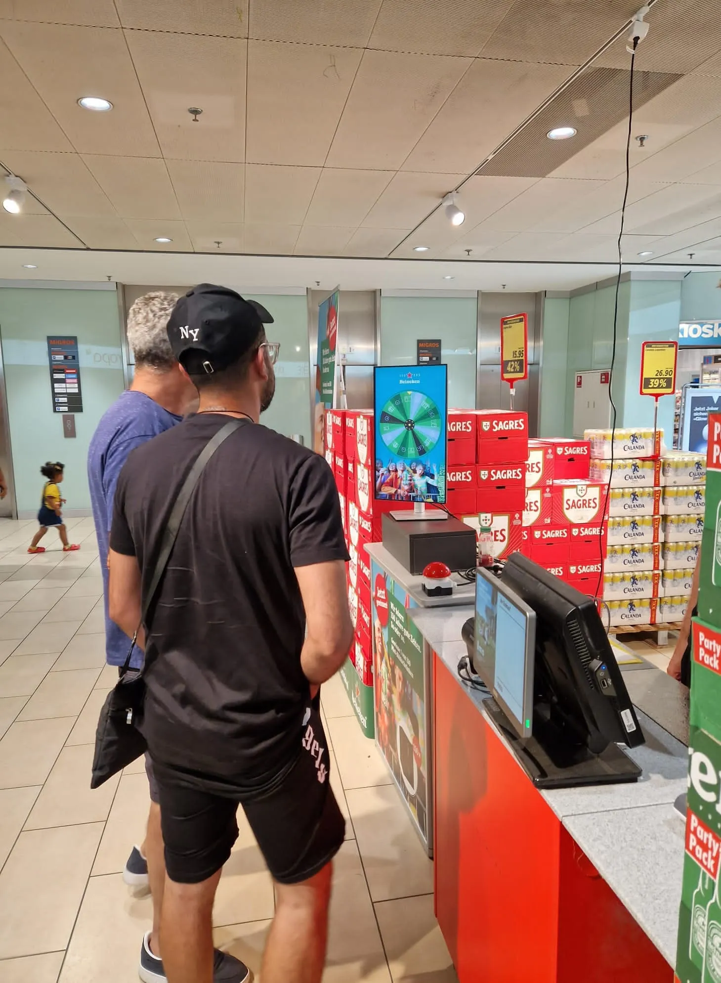 Zwei Männer vor der roten Theke mit Bildschirm des Heineken-Glücksrads, dahinter Sagres- und Calanda-Kisten im Laden