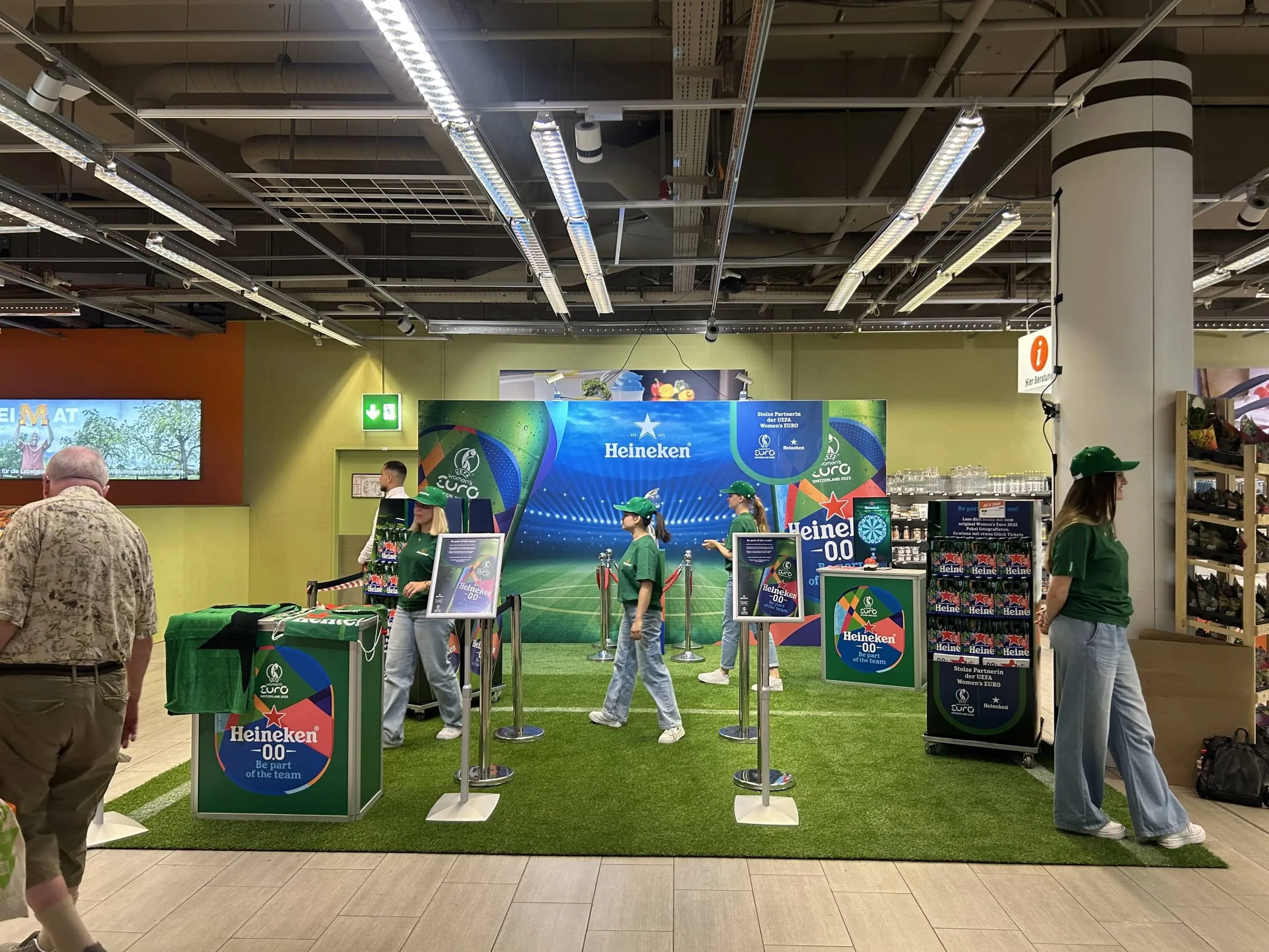 Heineken promotional area in Migros with artificial turf, a stadium photo backdrop, counters and staff in green T-shirts