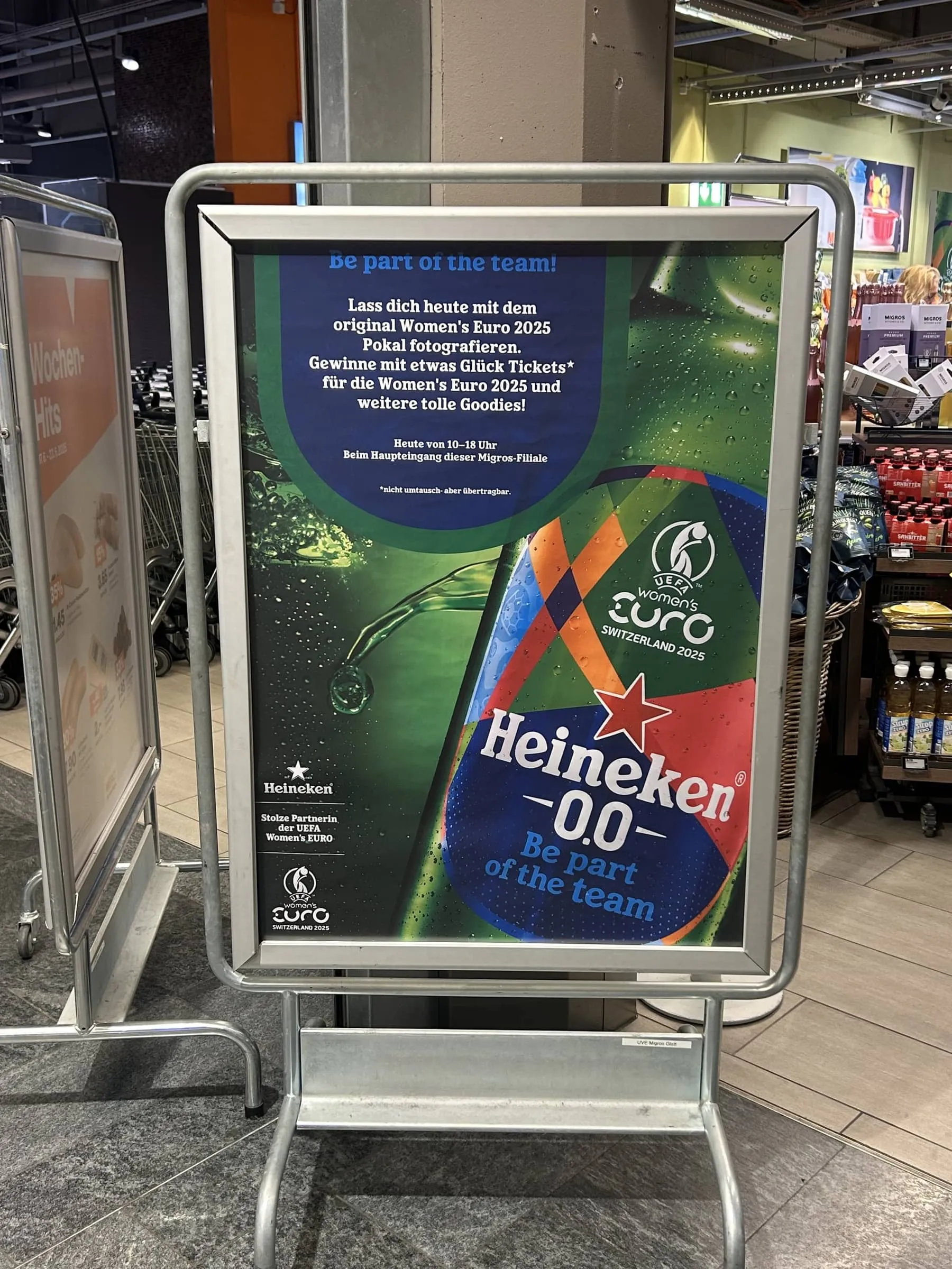 Plakatständer mit Heineken-0.0-Plakat «Be part of the team!» zum Pokal-Foto und Ticket-Gewinnspiel in der Migros