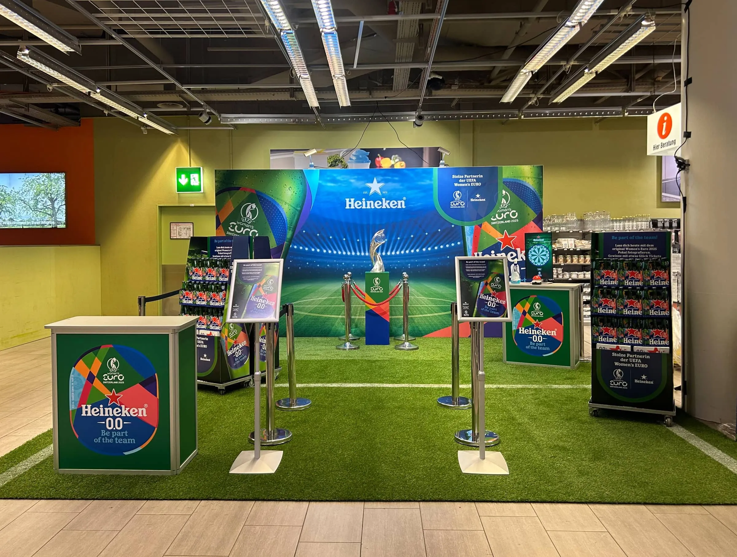 Heineken-Promotionsfläche mit Kunstrasen, Fotowand mit Stadionmotiv, Pokal auf Sockel hinter roter Kordel und Theken in der Migros