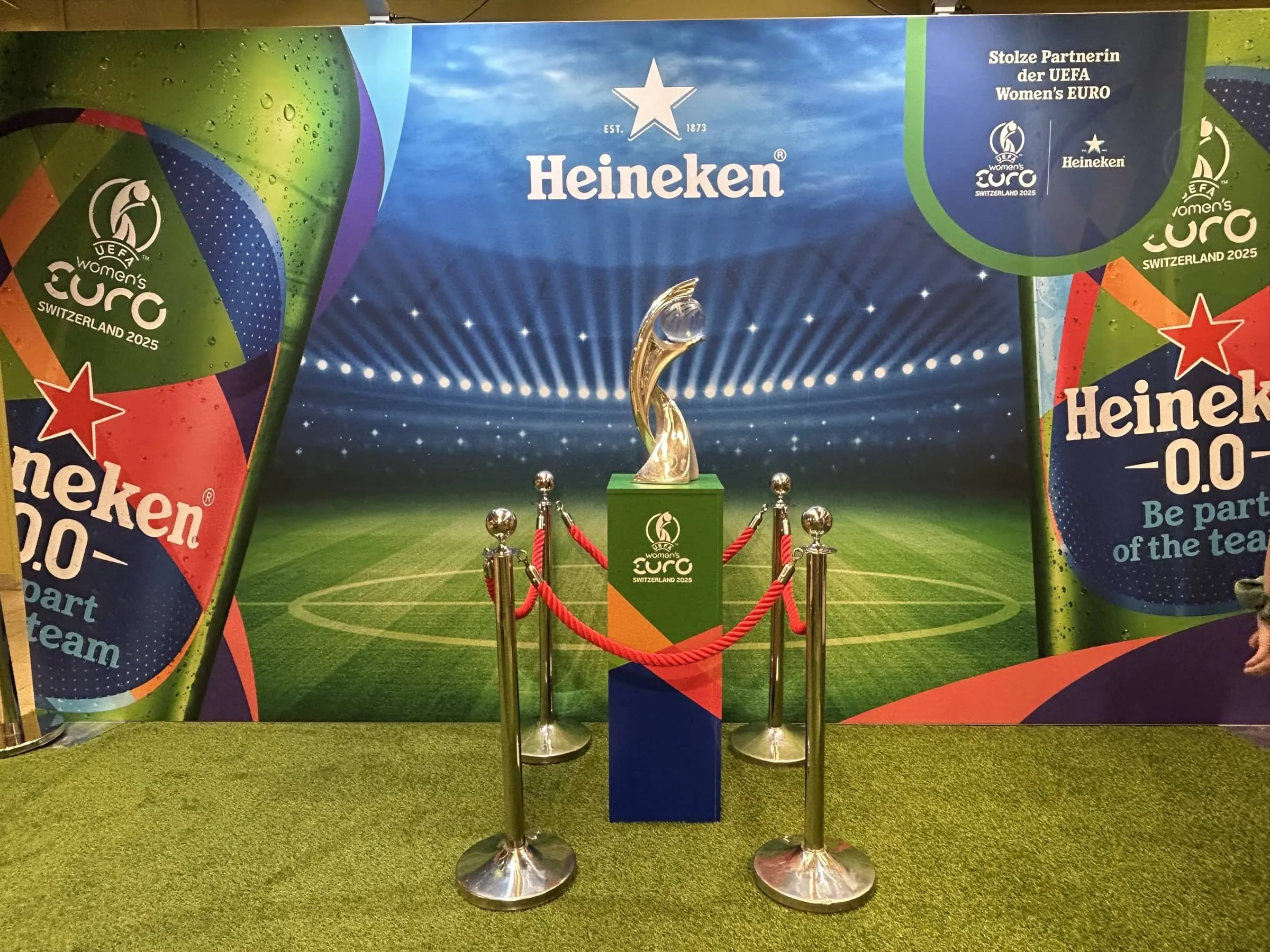 Silberner WEURO-Pokal auf farbigem Sockel hinter roter Kordelabsperrung vor der Heineken-Fotowand mit Stadionmotiv
