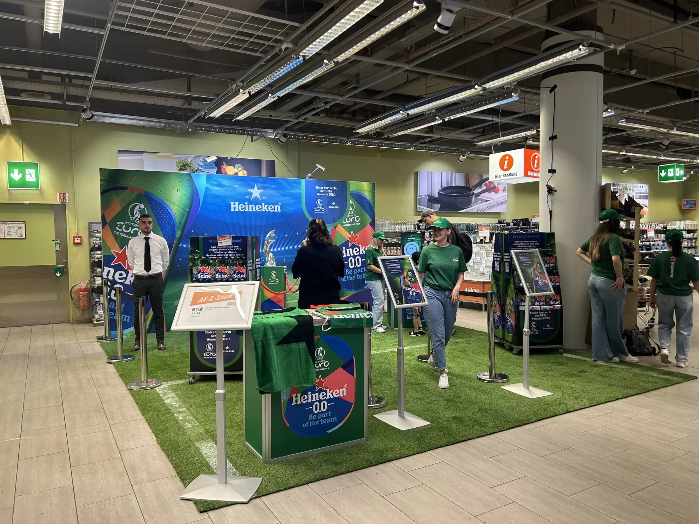 Promotorinnen in grünen Heineken-T-Shirts und Besucher auf dem Kunstrasen, Theke mit Heineken-0.0-Branding im Vordergrund