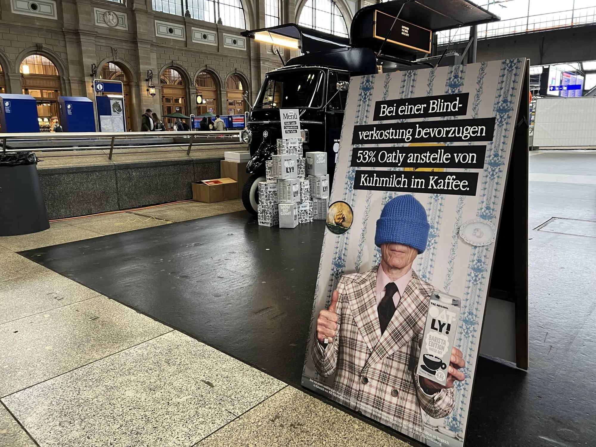 Kundenstopper mit Oatly-Plakat zur Blindverkostung vor schwarzem Kaffee-Piaggio und gestapelten Oatly-Kartons in der Bahnhofshalle