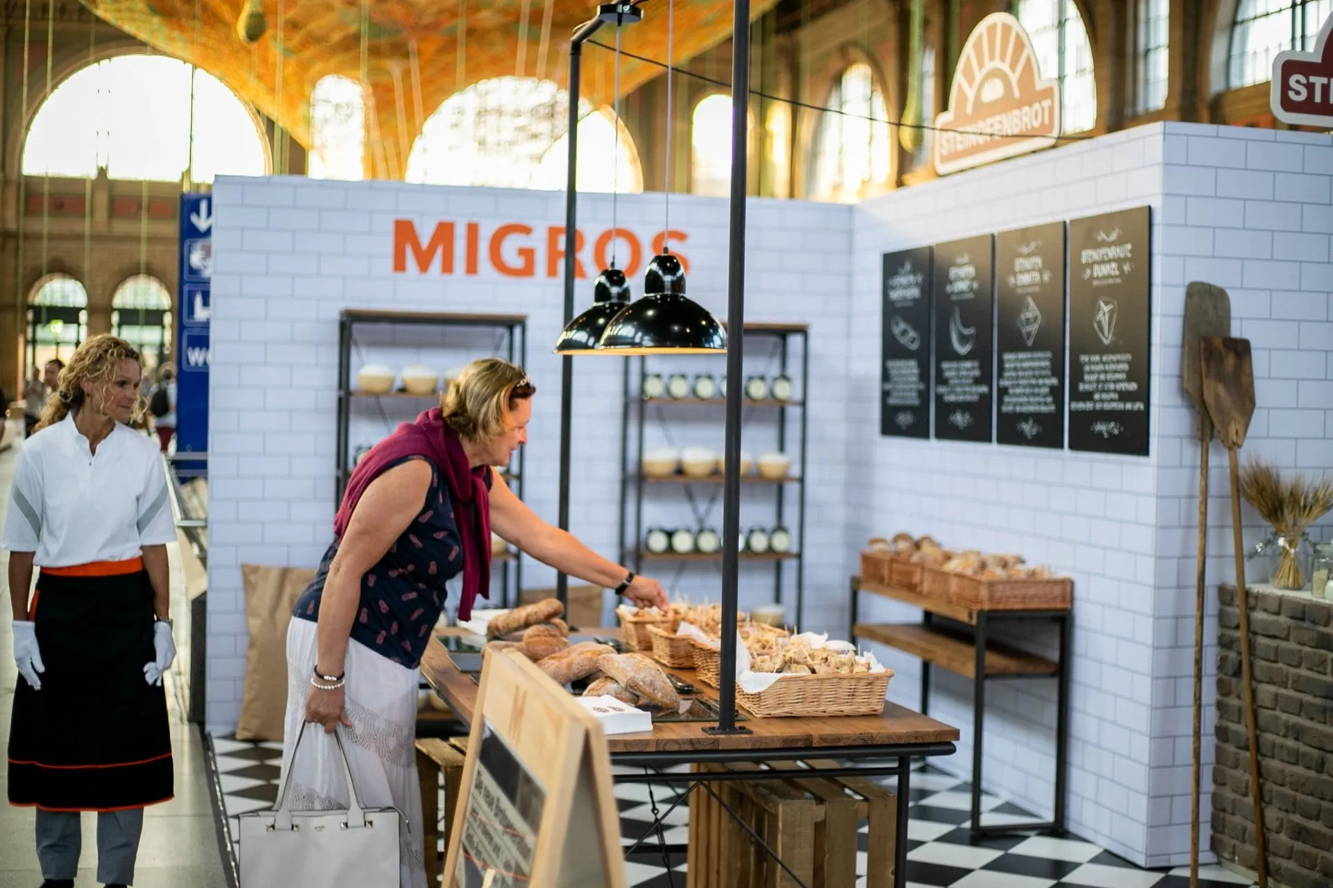 Besucherin greift nach Brothäppchen am Stand mit weisser Kachelwand und Migros-Schriftzug