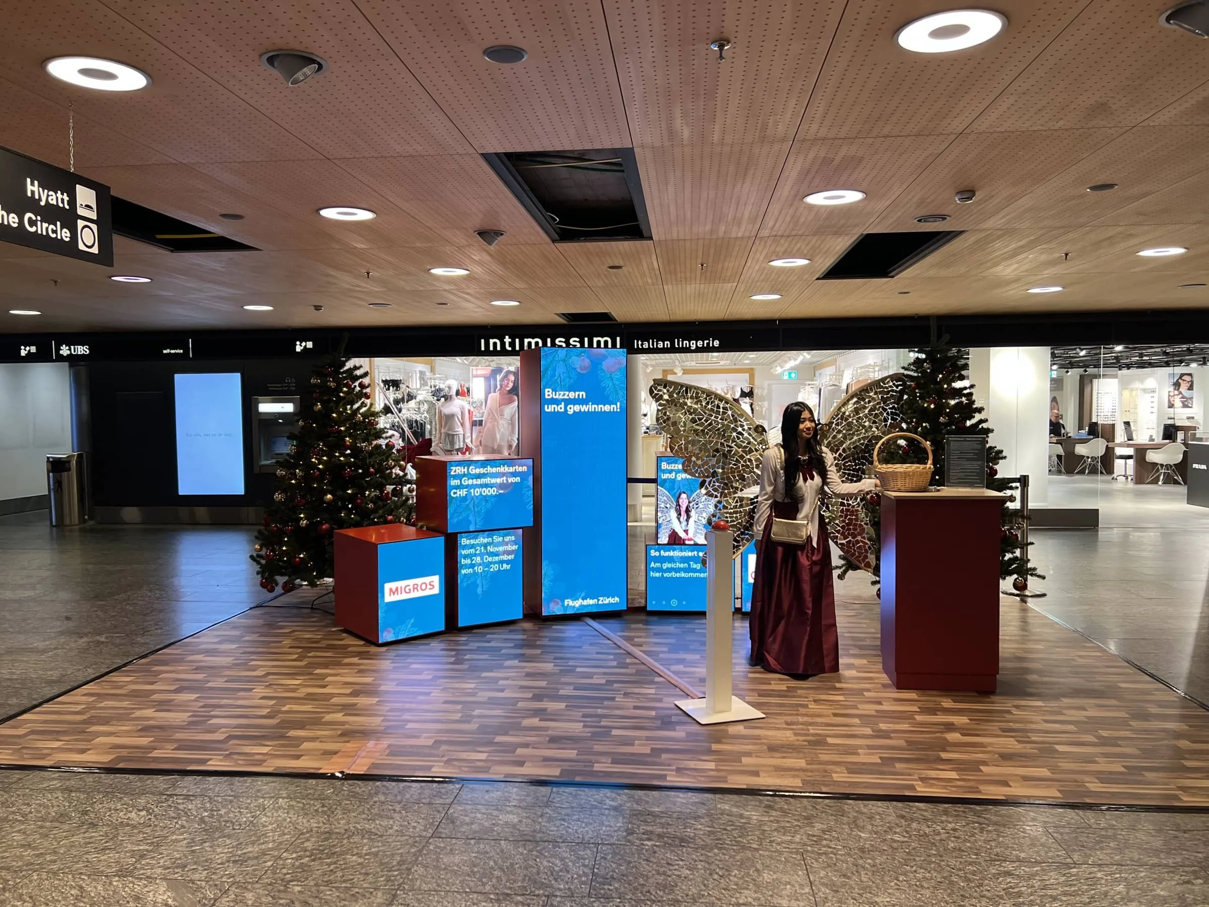 Christmas fairy with gold mirrored wings beside a red buzzer pedestal, blue «Buzzern und gewinnen!» screens and Christmas trees in Airport Shopping
