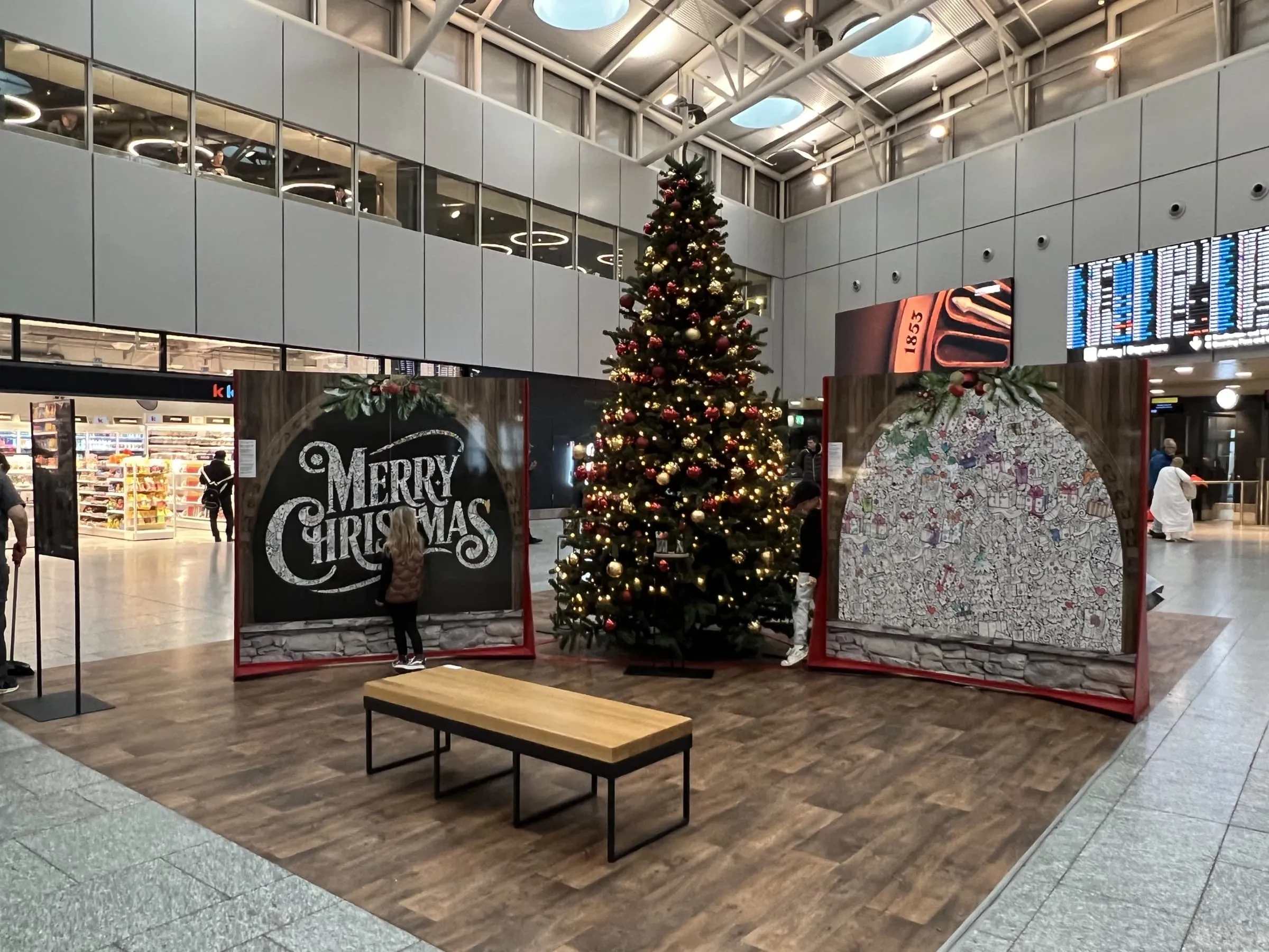 Two walls with «Merry Christmas» lettering and a mandala colouring design beside a decorated airport Christmas tree, with a child colouring