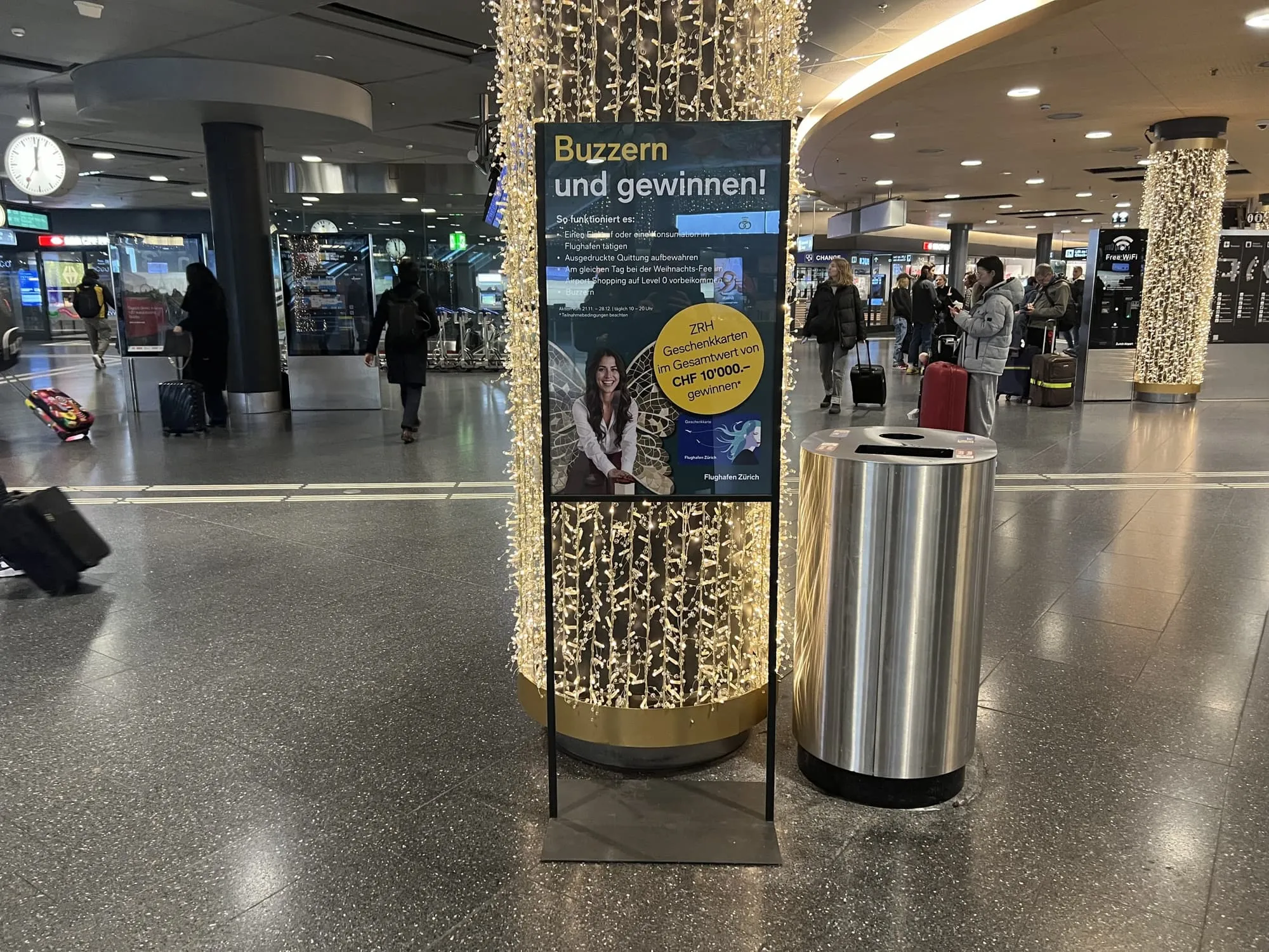 Plakatstele «Buzzern und gewinnen!» an einer Lichterketten-Säule im Flughafen, Reisende mit Rollkoffern im Hintergrund