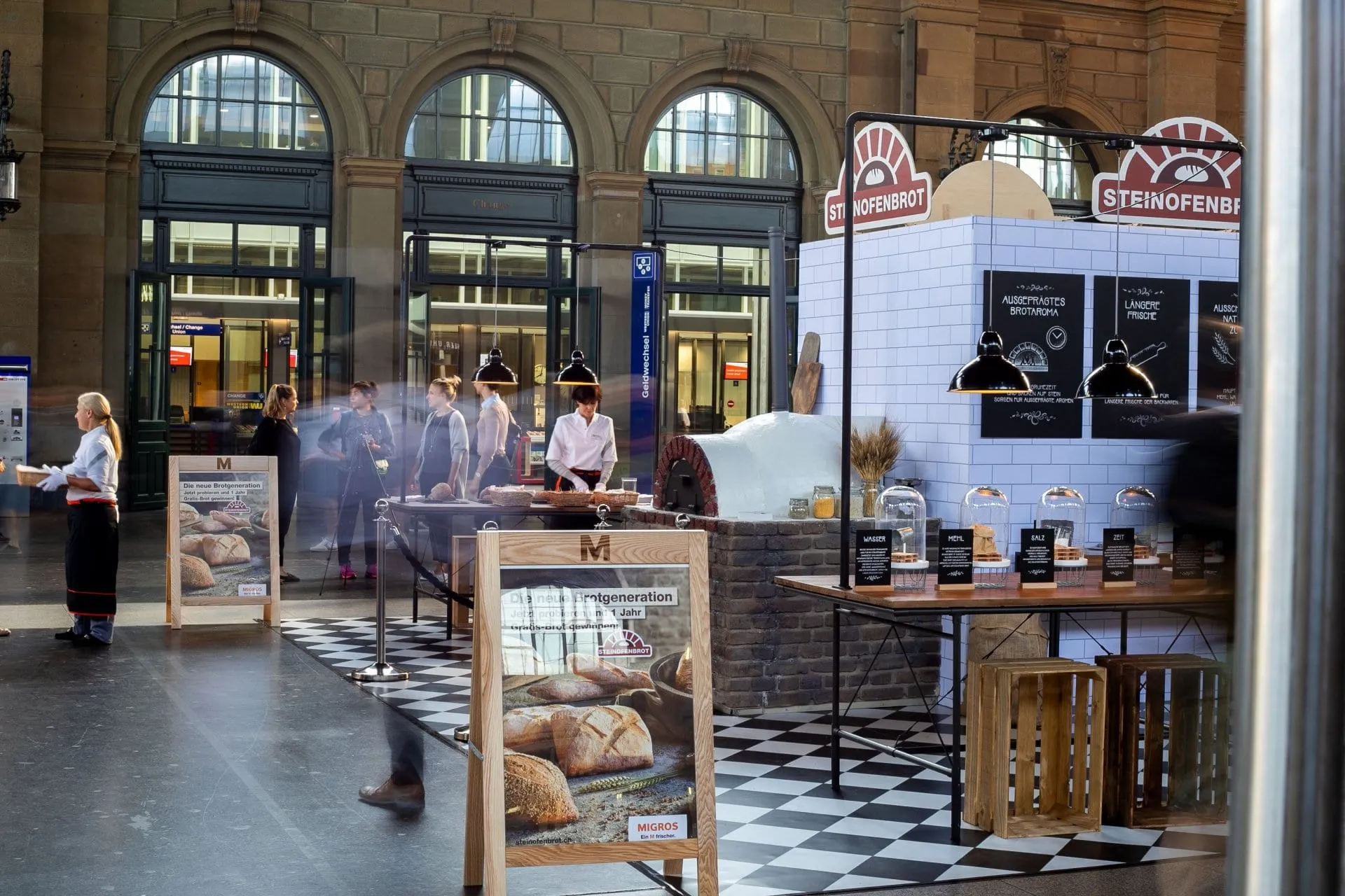Stand mit Steinofen, Tafeln und Glasglocken mit Zutaten, Promotorin und Besuchende im Hintergrund