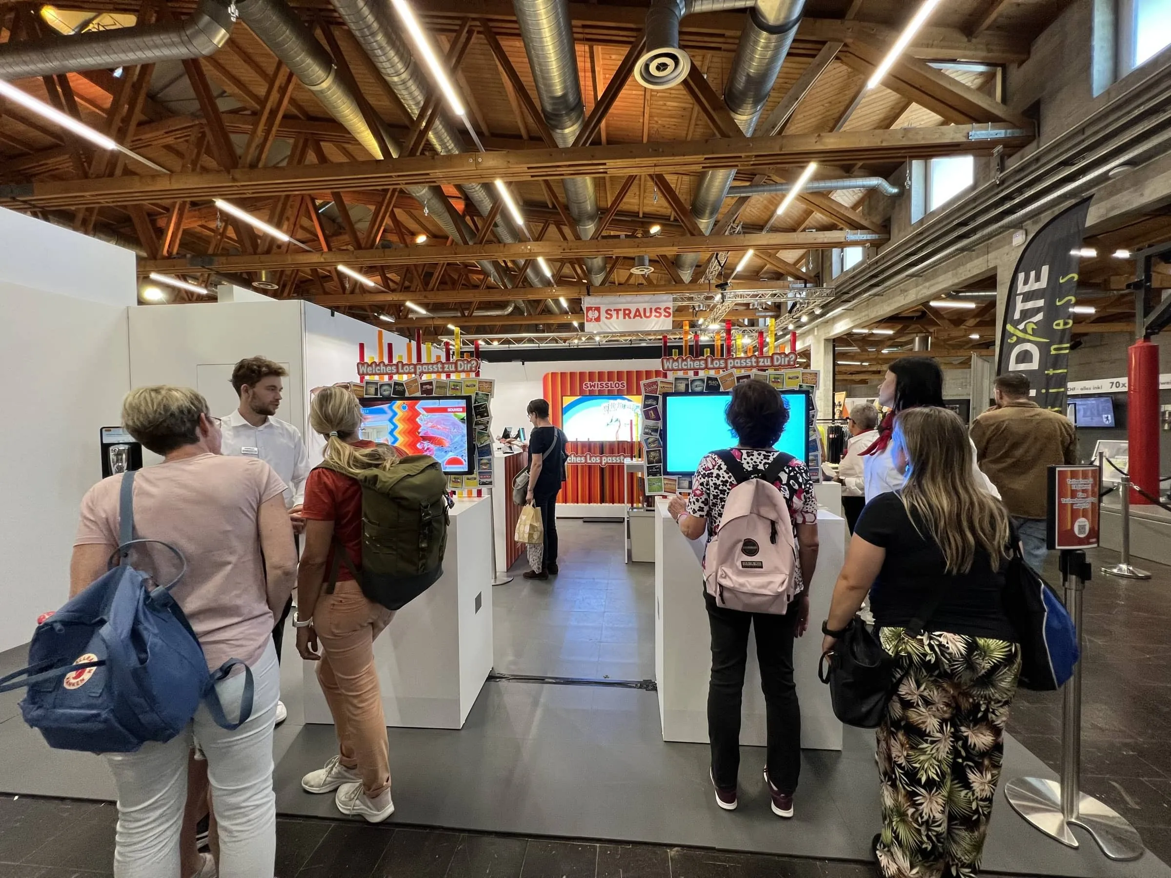 Besuchende an den beiden Game-Stationen «Welches Los passt zu Dir?» in einer Halle mit Holzdach