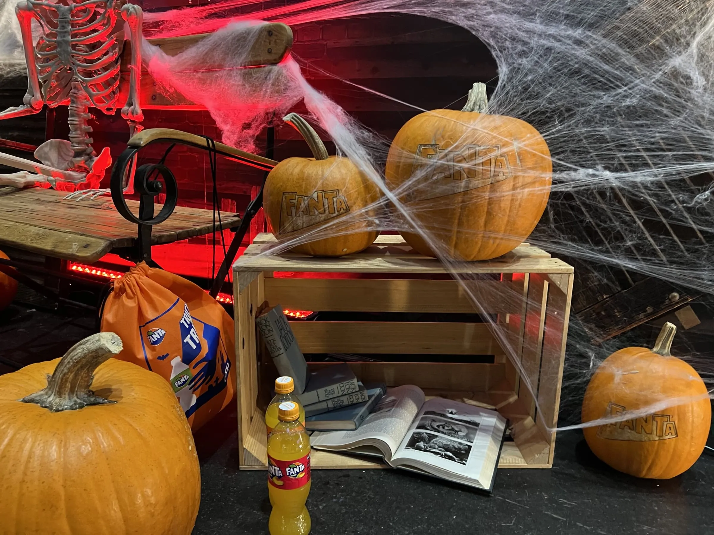 Kürbisse mit eingraviertem Fanta-Logo auf einer Holzkiste, dazu Skelett, Spinnweben, Fanta-Flasche und alte Bücher