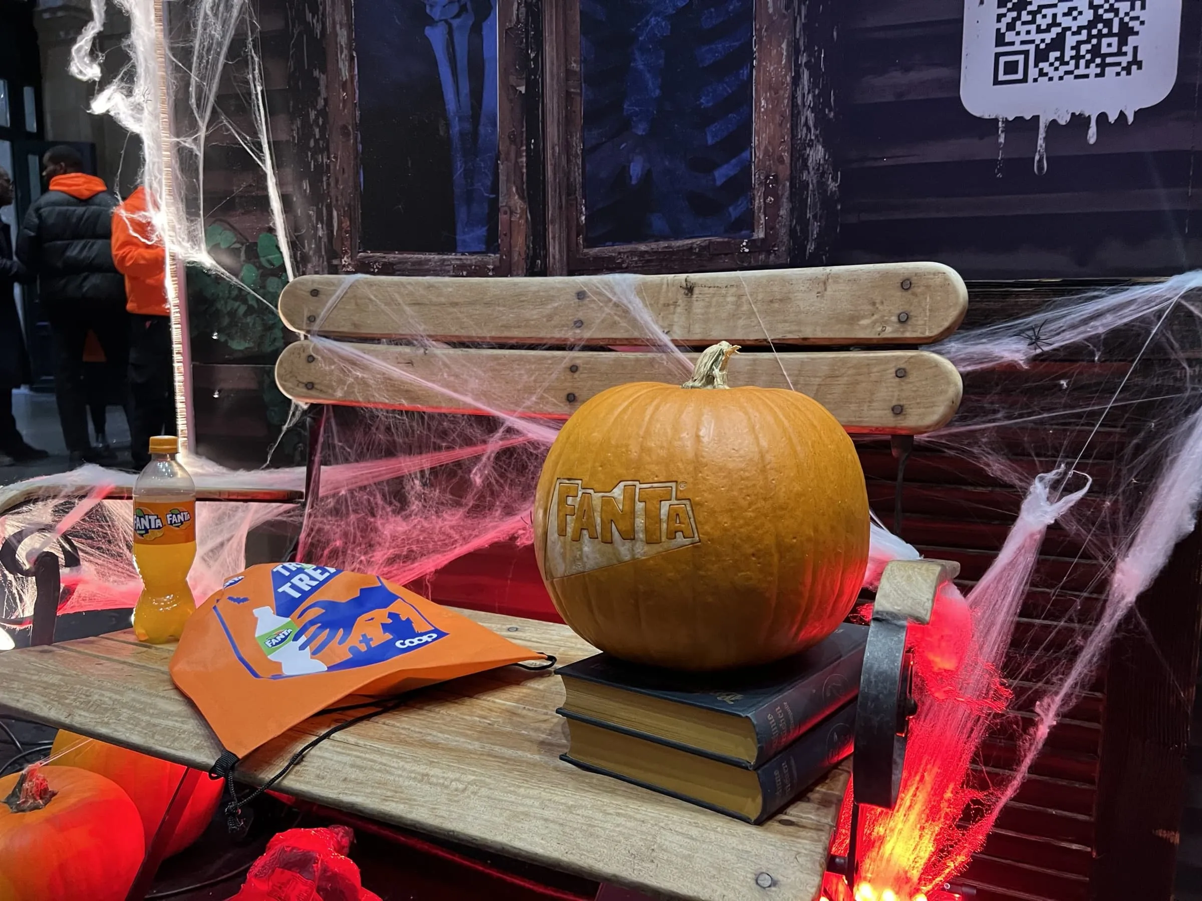 Holzbank mit Fanta-Kürbis auf Büchern, orangem Fanta-Beutel, Flasche, Spinnweben und QR-Code