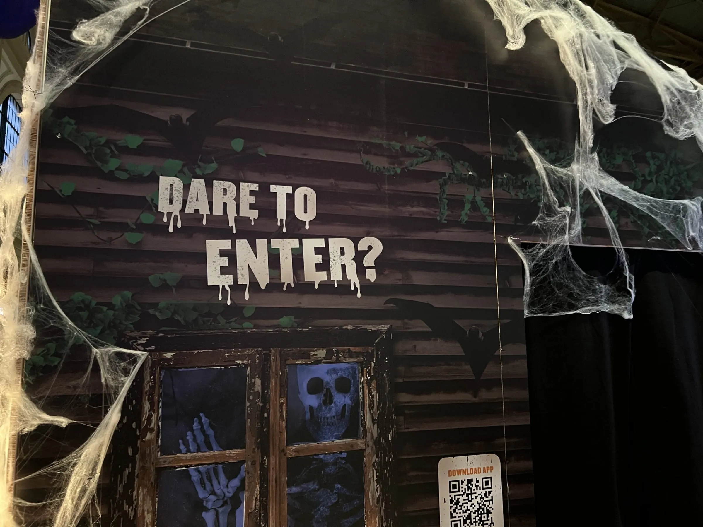 Holzwand mit Schriftzug «Dare to Enter?», Fledermäusen, Skelett im Fenster und QR-Code «Download App»