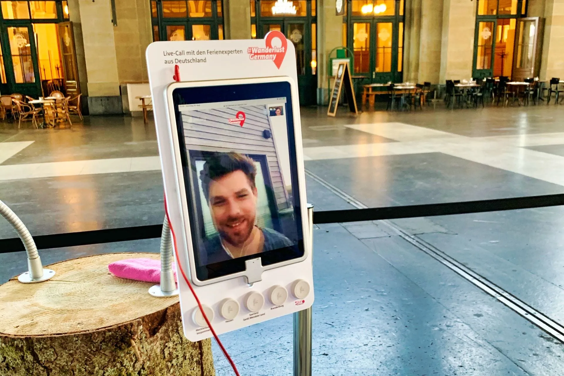 Videocall-Station auf einem Baumstamm: Tablet mit lächelndem Mann, Schild «Live-Call mit den Ferienexperten aus Deutschland»