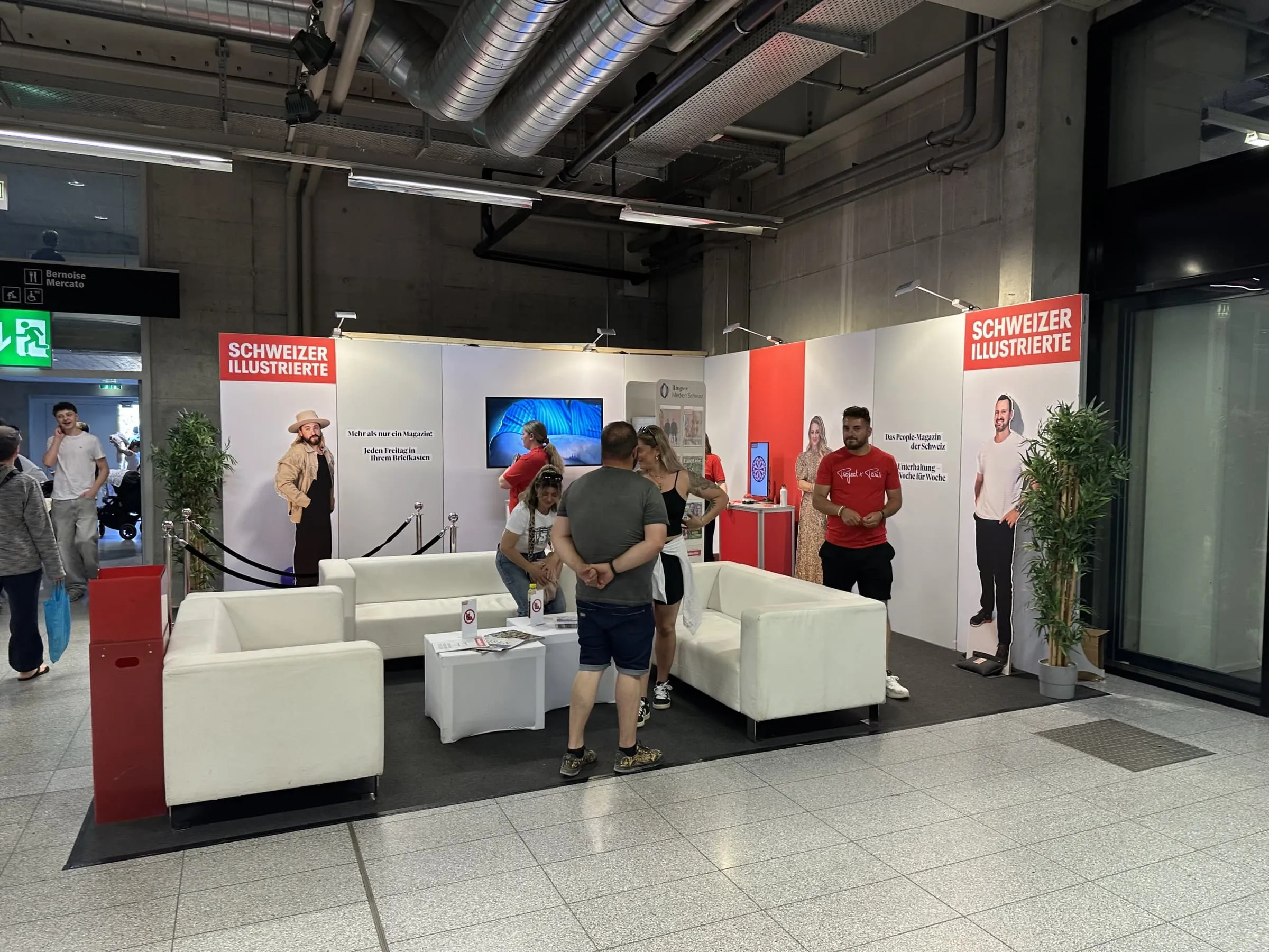 Offener SI-Eckstand mit weissen Sofas, roter Wandfläche, Star-Cutouts und Promotor in roten Shirts im Gespräch mit Besuchenden