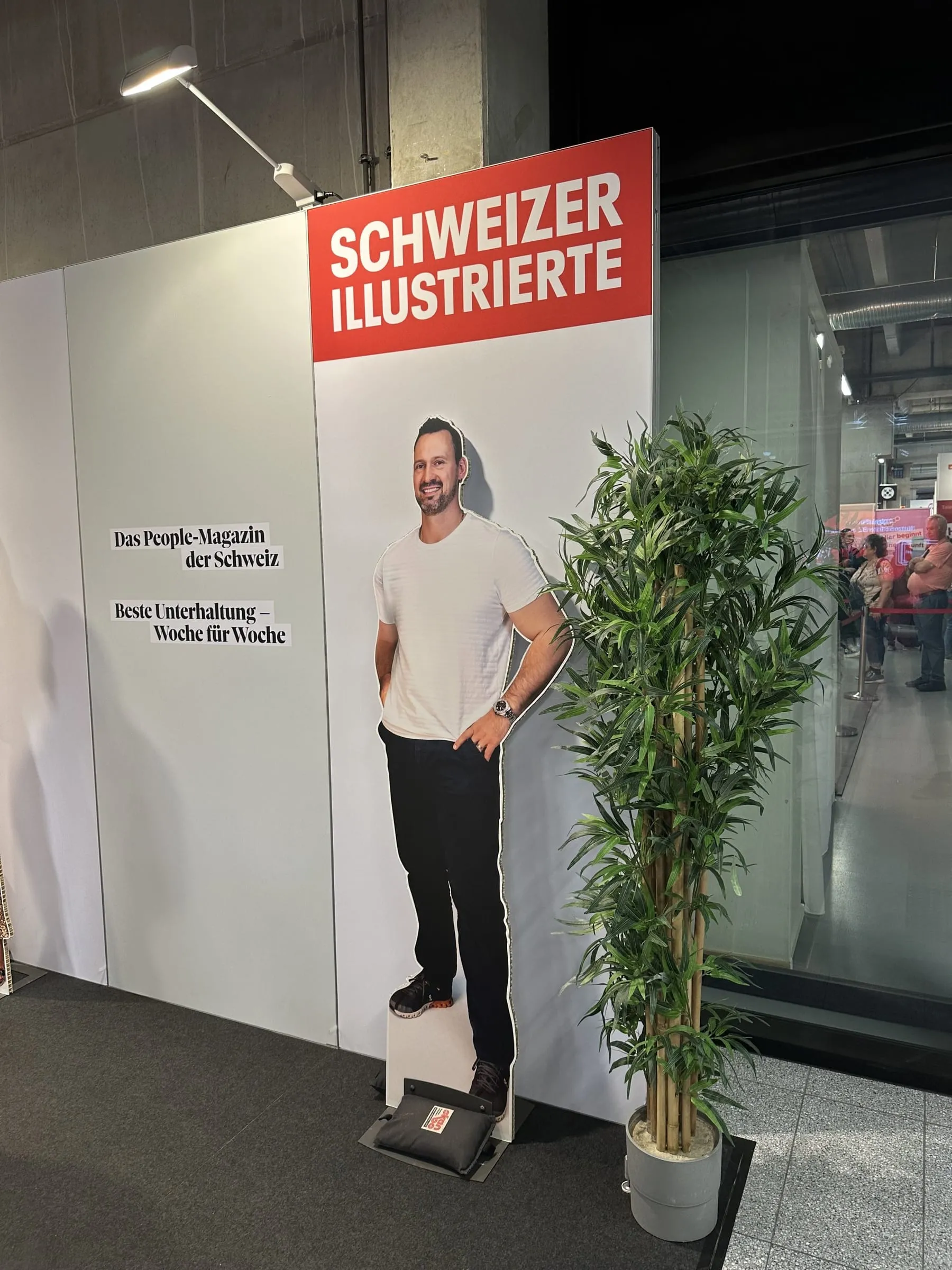 Lebensgrosses Cutout eines Mannes im weissen T-Shirt neben dem SI-Logo und einer Bambuspflanze