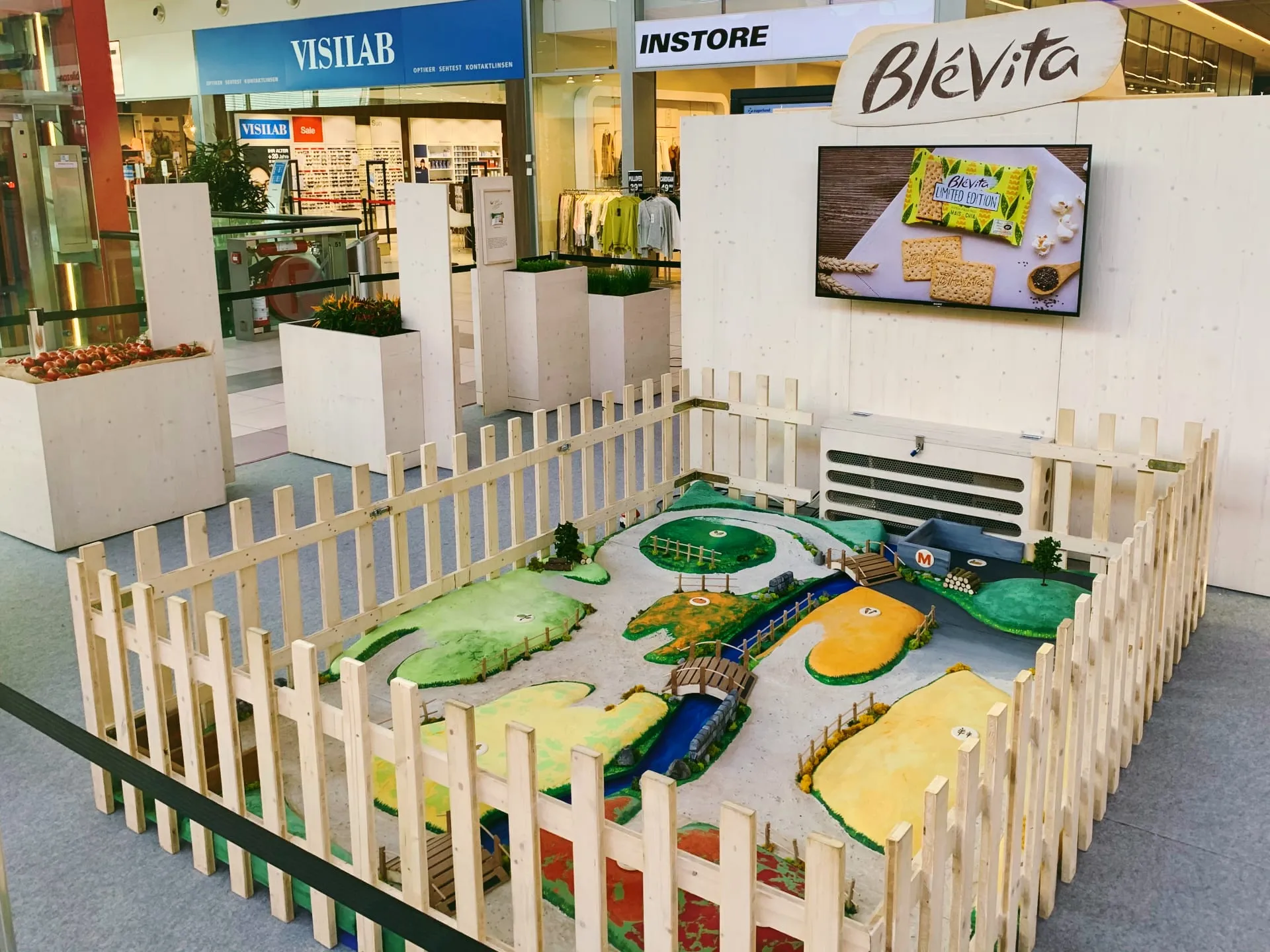 Blévita stand in a shopping centre with a fenced miniature landscape of colourful fields, a river and a screen