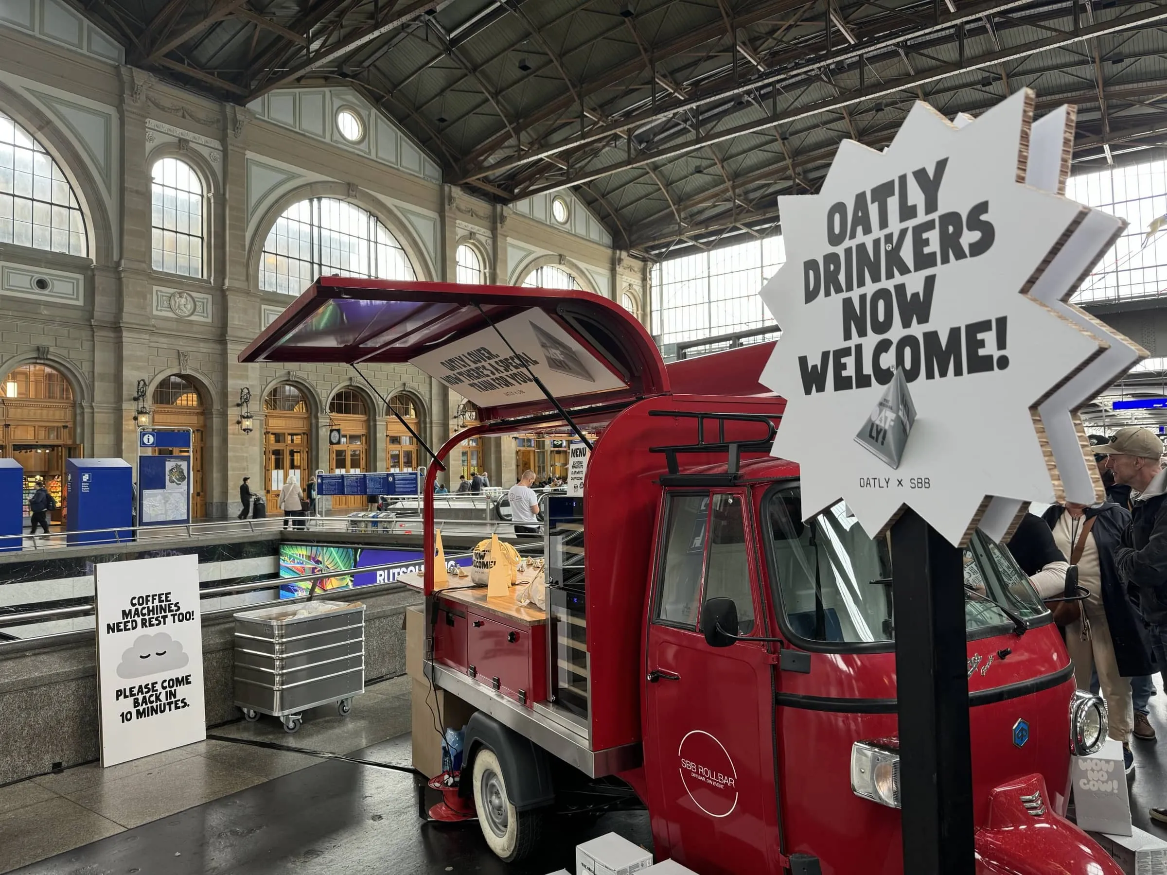 Red Piaggio Ape «SBB Rollbar» with an «Oatly Drinkers Now Welcome!» sign in the hall of Zürich main station