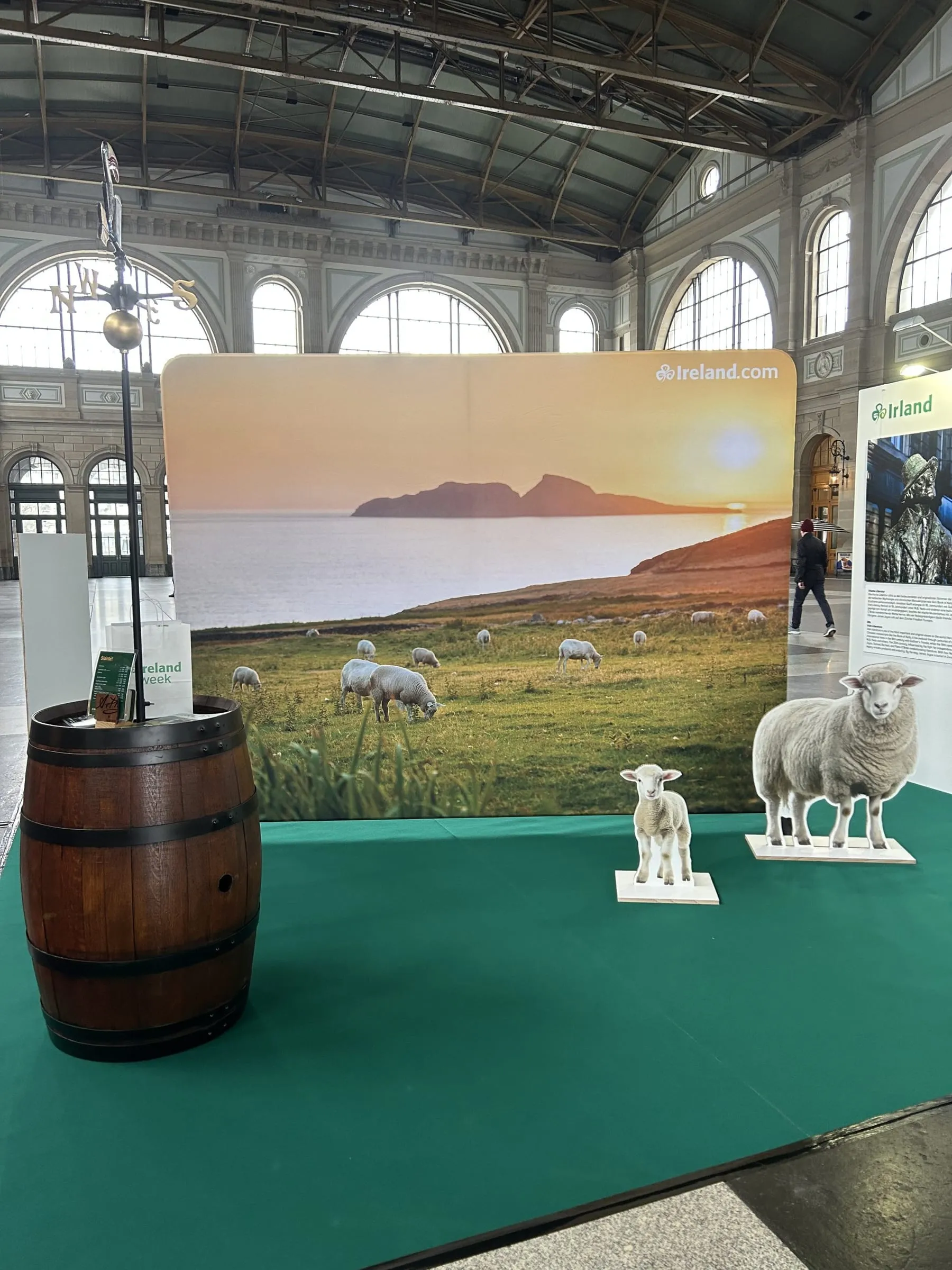 Fotowand mit Schafen im Sonnenuntergang, zwei Pappschafe und Holzfass mit Wetterfahne auf grünem Teppich in der Bahnhofshalle