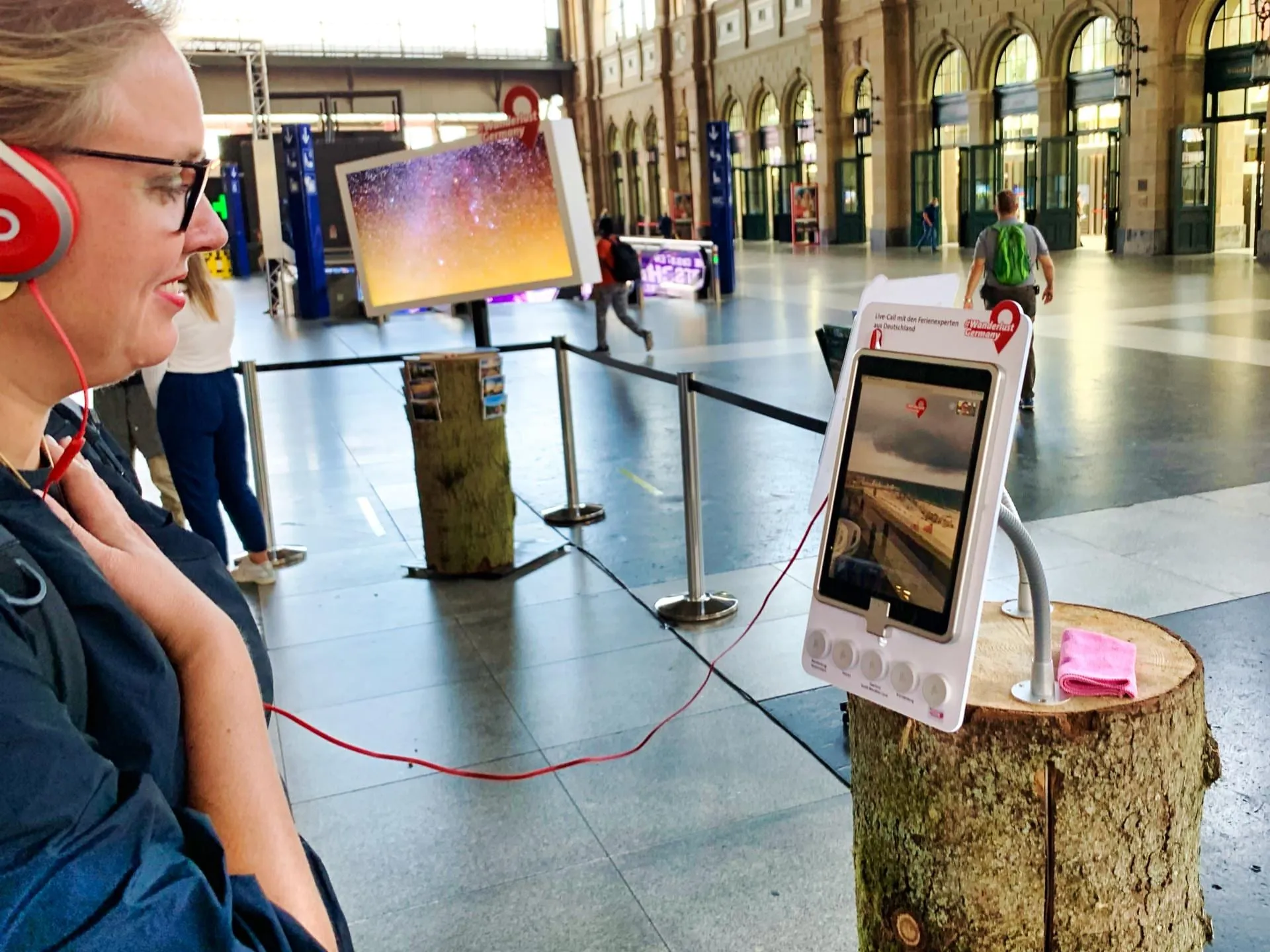 Besucherin mit roten Kopfhörern an der Videocall-Station, Tablet zeigt einen Strand, dahinter die Bahnhofshalle