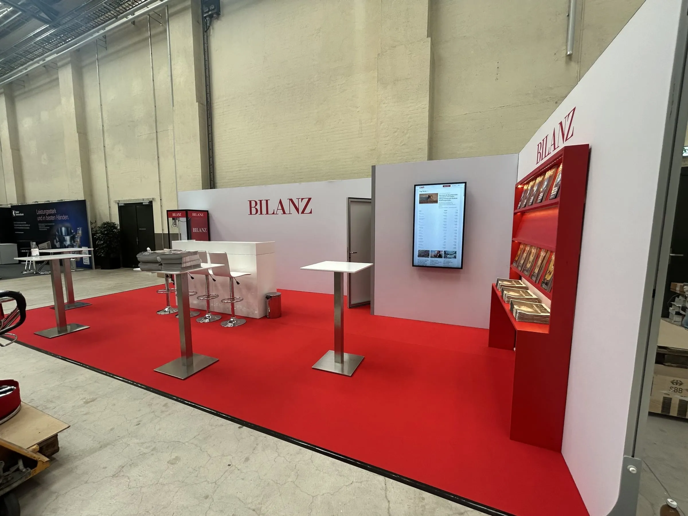 Stand von der Seite mit weisser Bar, Barhockern, Bildschirm mit Cash-Newsseite und rotem BILANZ-Regal an der Rückwand