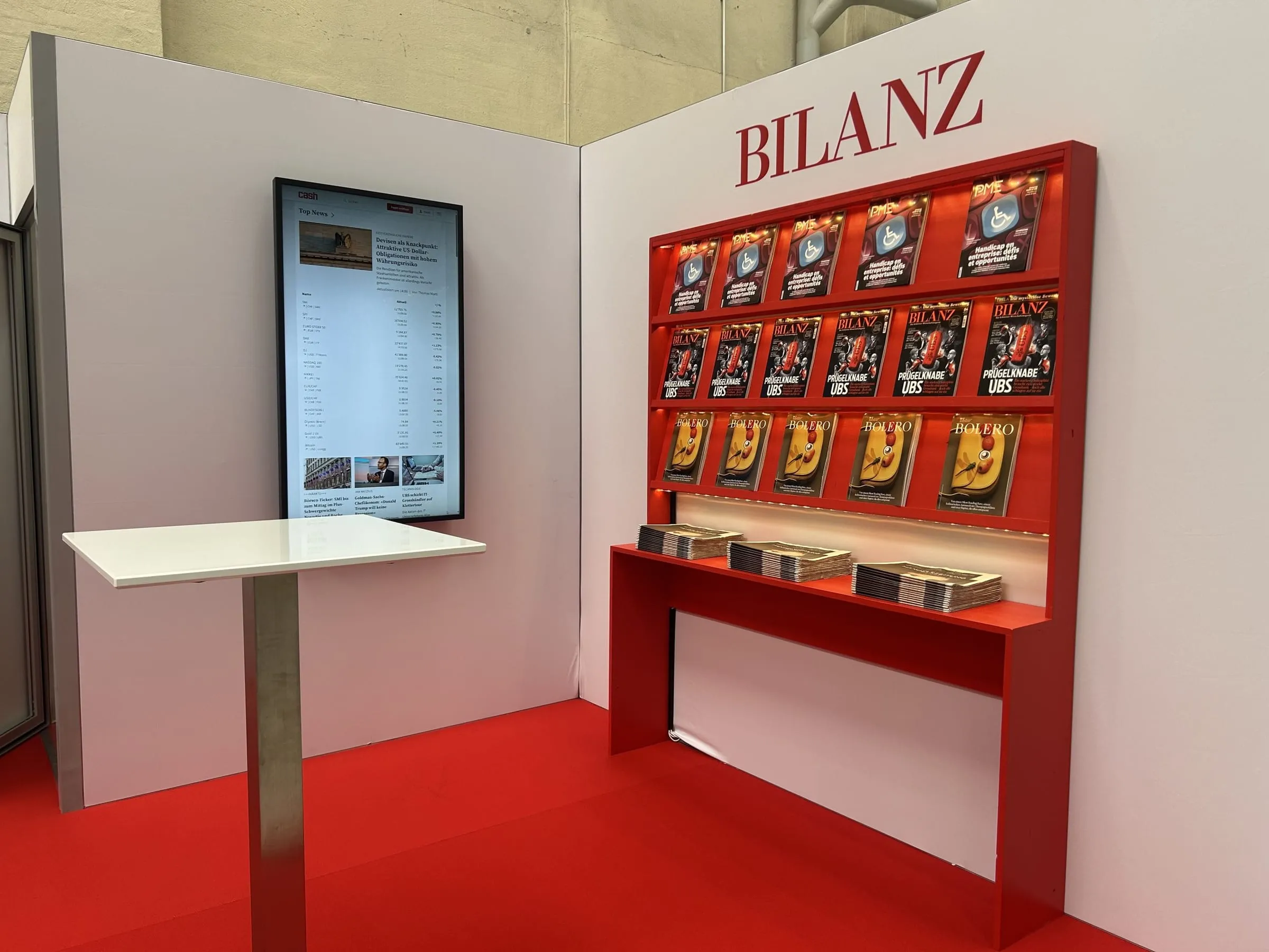 Red magazine-shelf area of the BILANZ stand with a screen, standing table and magazines on a red carpet