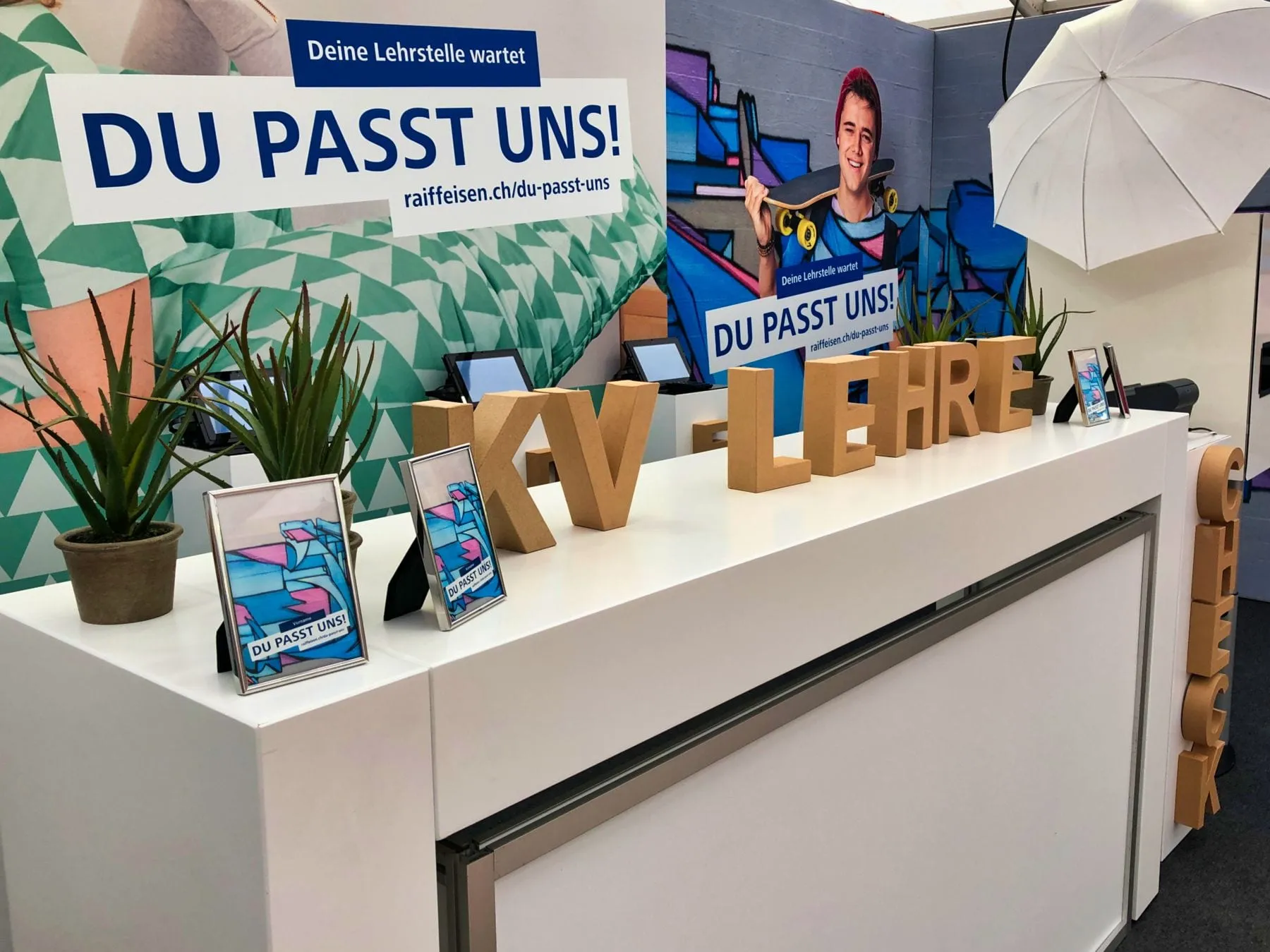 White counter with cardboard «KV-Lehre» letters, tablets and framed photos in front of Raiffeisen «Du passt uns!» posters
