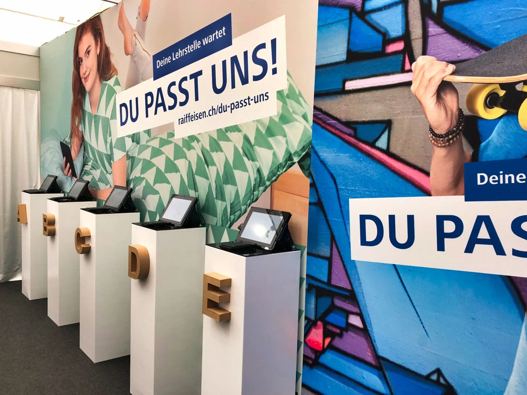 Fünf weisse Stelen mit Tablets und Buchstaben A bis E für den Typentest vor Raiffeisen-Kampagnenwänden