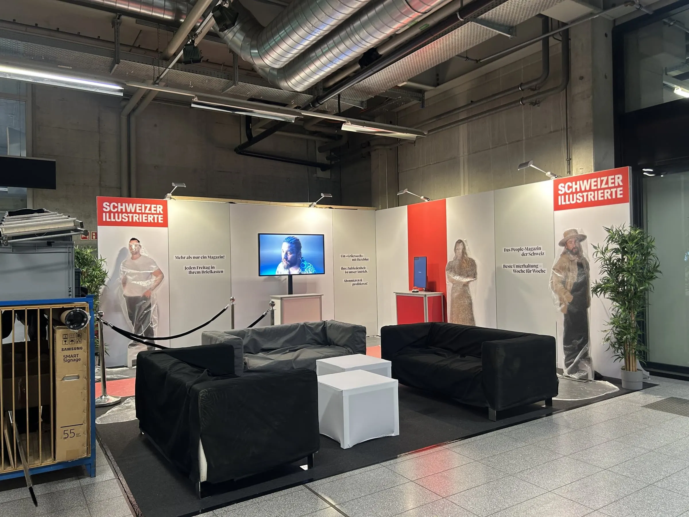 Fertiger Stand vor der Eröffnung: Sofas noch abgedeckt, Star-Cutouts in Folie, Bildschirm und Kordelabsperrung