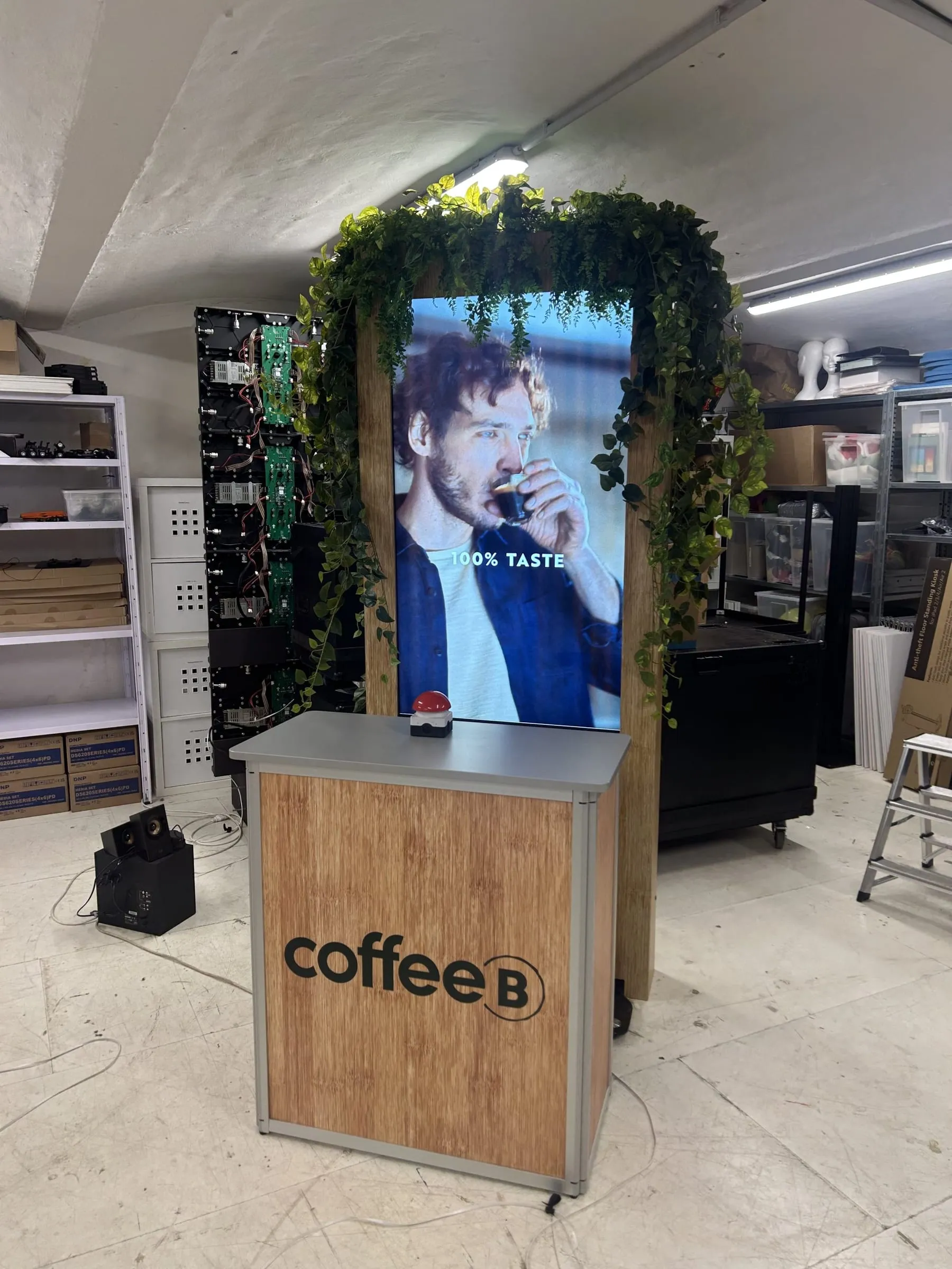 Counter mit Coffee-B-Logo und rotem Buzzer vor dem Screen, der einen Mann beim Espressotrinken mit «100% Taste» zeigt