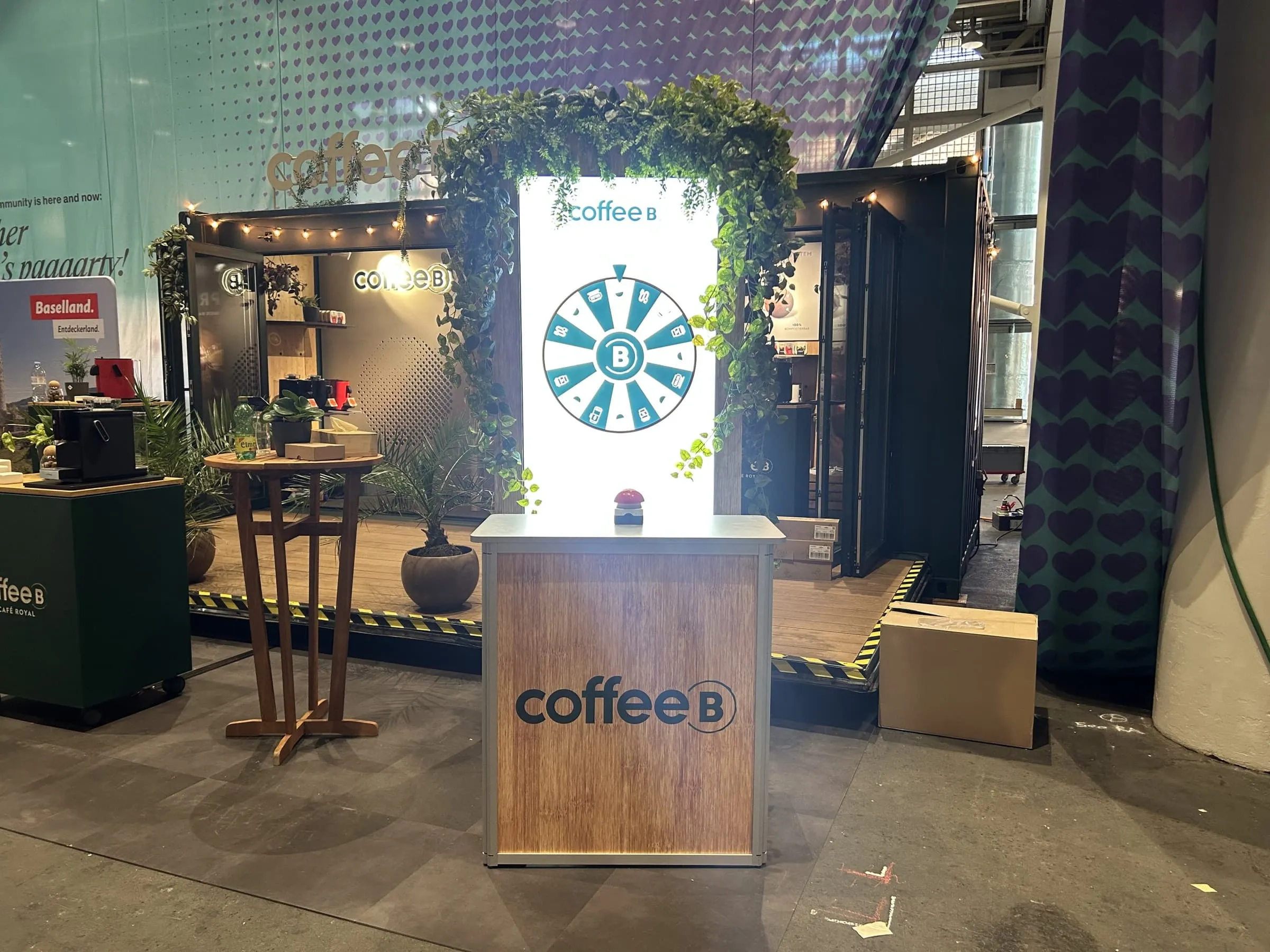 Glücksrad-Counter mit Coffee-B-Logo und rotem Buzzer, dahinter der Screen mit Glücksrad im bepflanzten Holzrahmen vor dem Container
