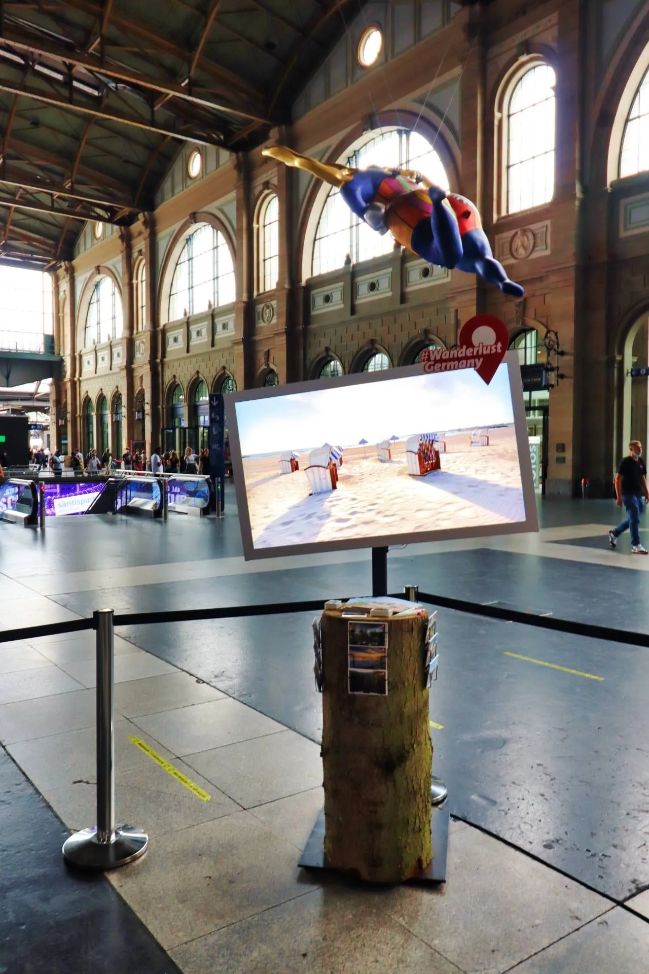 Bildschirm mit Strandkörben auf einem Baumstamm, darüber die schwebende Figur in der Bahnhofshalle Zürich