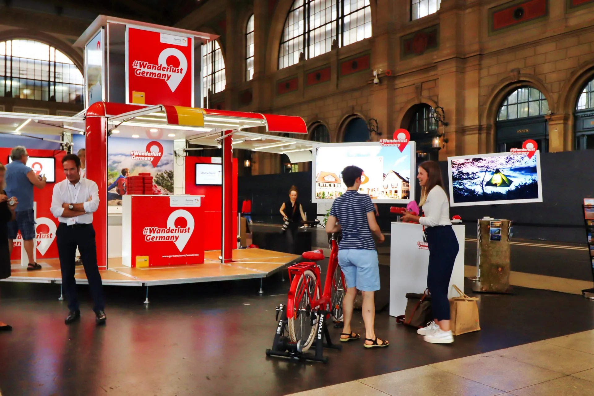 Roter Cube mit #Wanderlust Germany, rotes Velo auf der Rolle, Besucherinnen an einem Bildschirm mit Ferienbildern