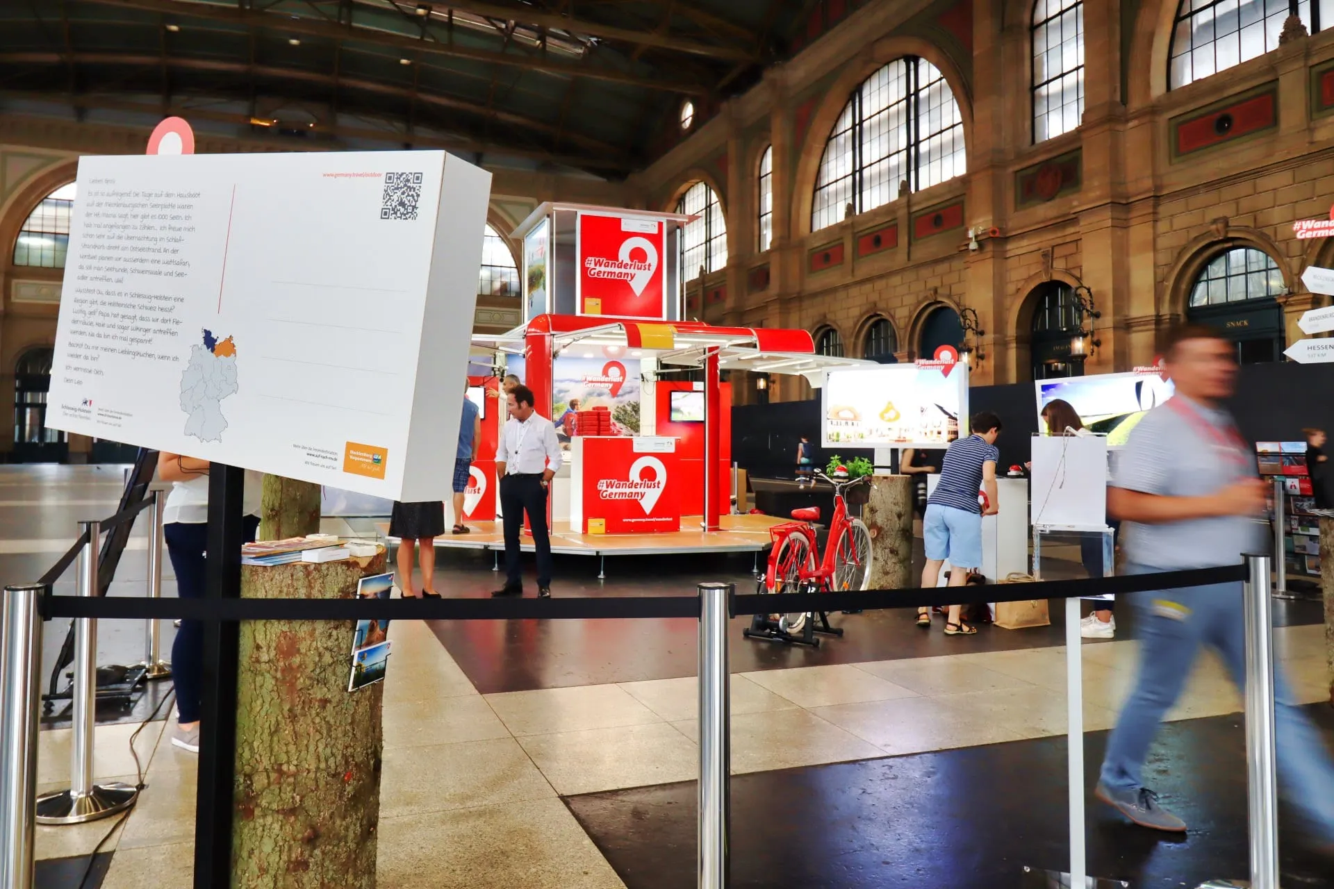 Riesenpostkarte im Vordergrund, dahinter roter Cube, rotes Velo und Besuchende in der Bahnhofshalle