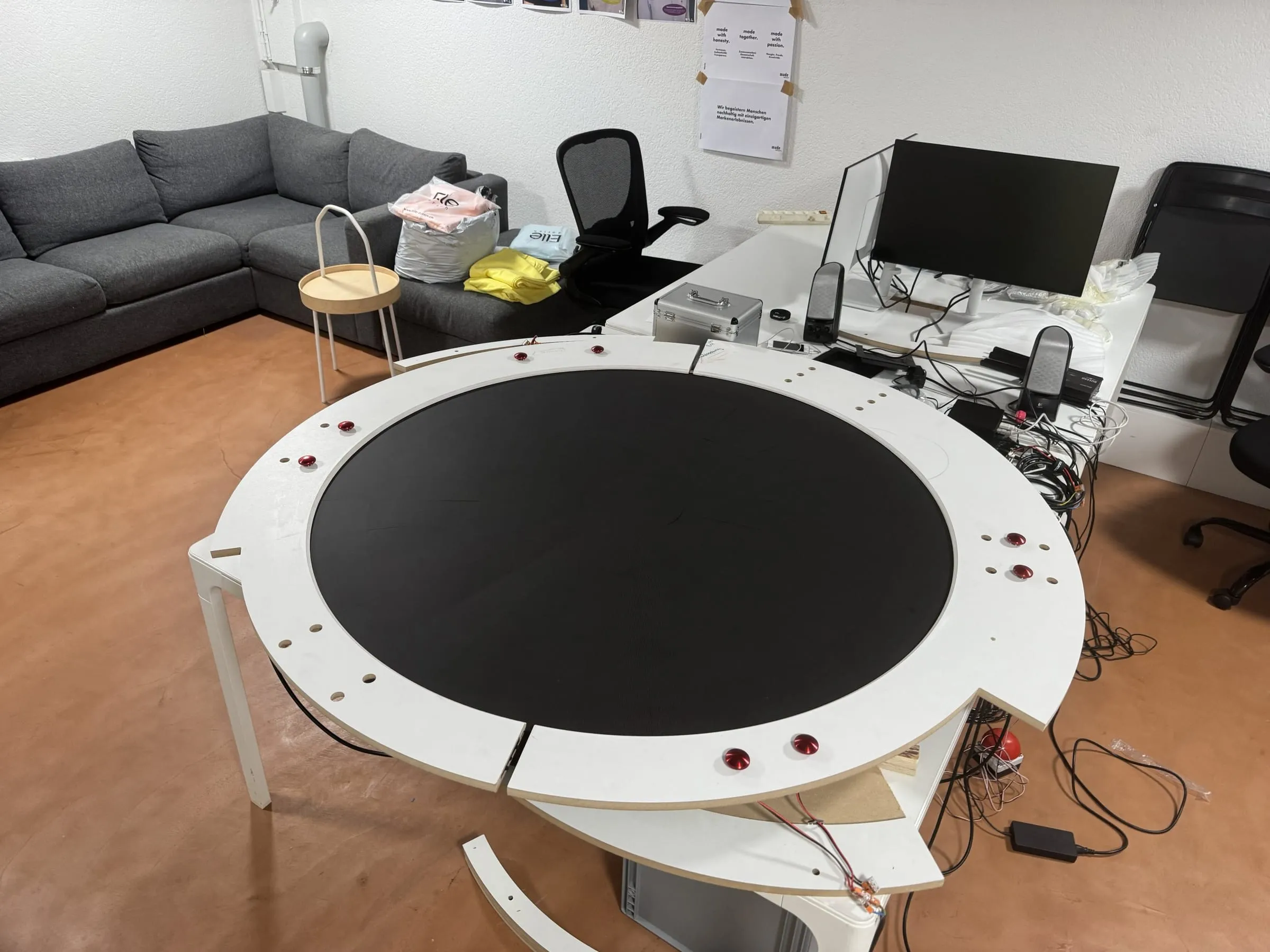Weisser runder Tischrahmen mit schwarzem LED-Screen und roten Buttons im Büro, daneben Kabel und ein Monitor