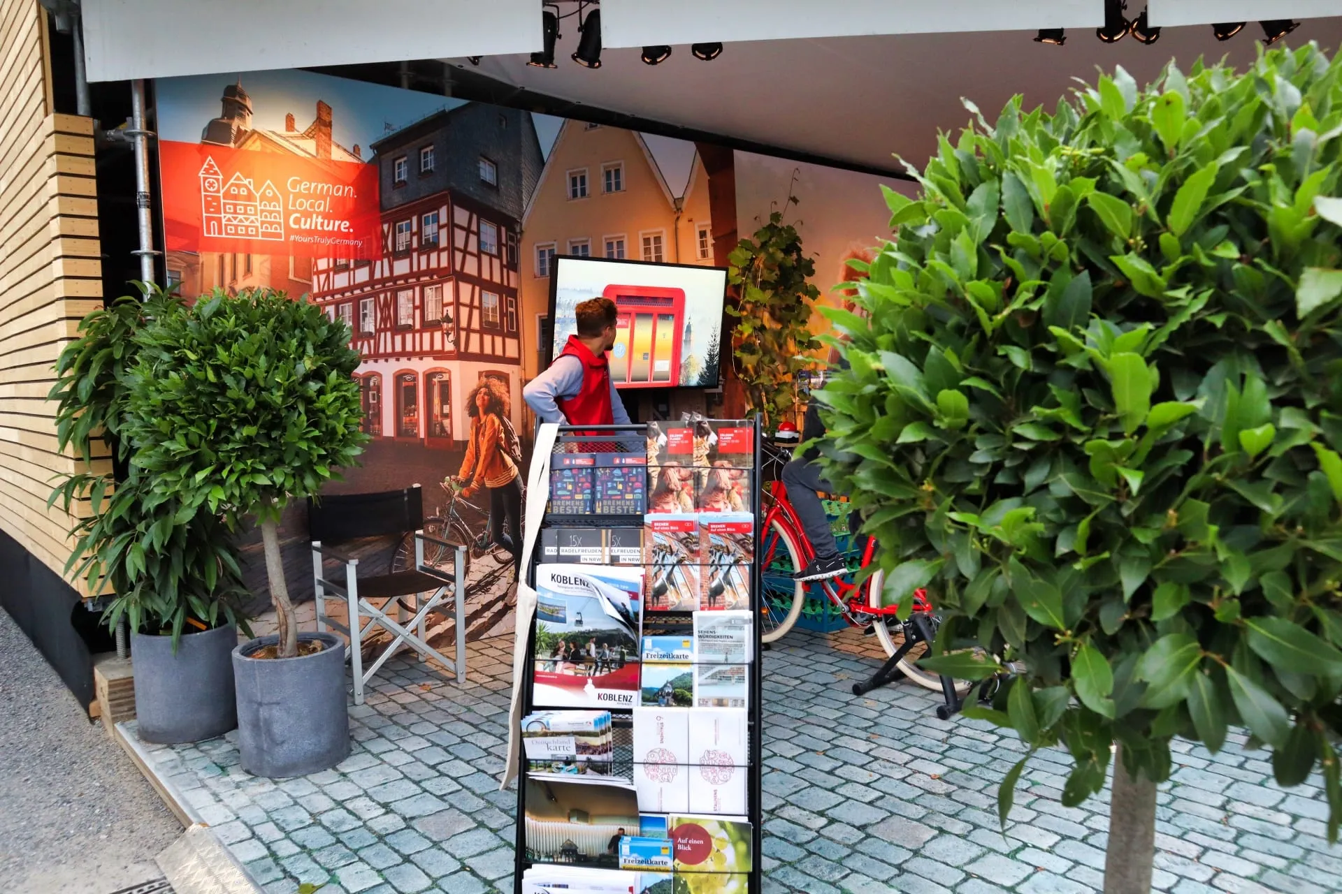 Prospektständer mit Reiseführern vor Fotowand mit Fachwerkhäusern, Bildschirm und Lorbeerbäumchen am Stand
