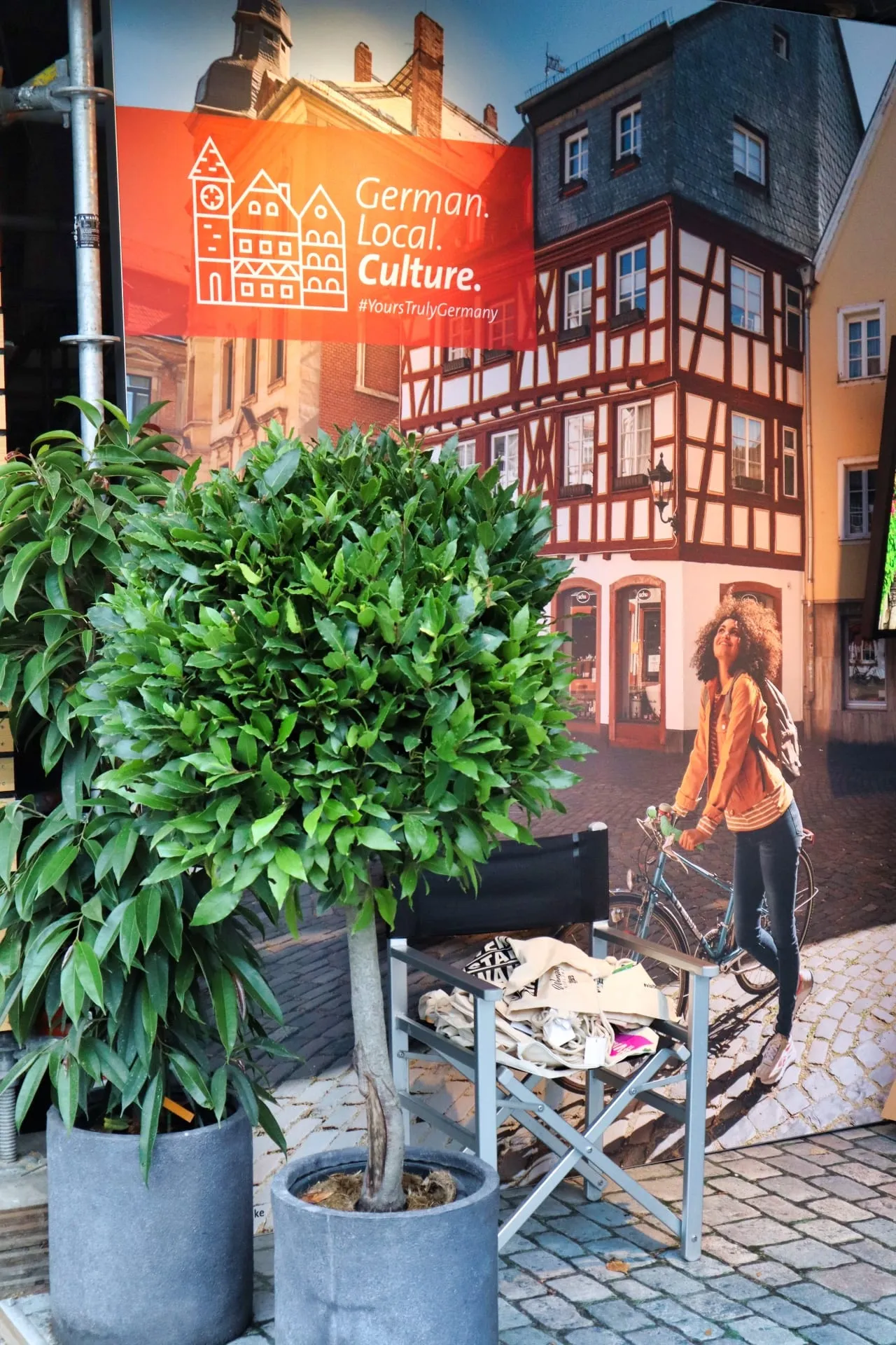 Lorbeerbäumchen und Regiestuhl mit Stofftaschen vor der Fotowand «German. Local. Culture.» mit Fachwerkhaus
